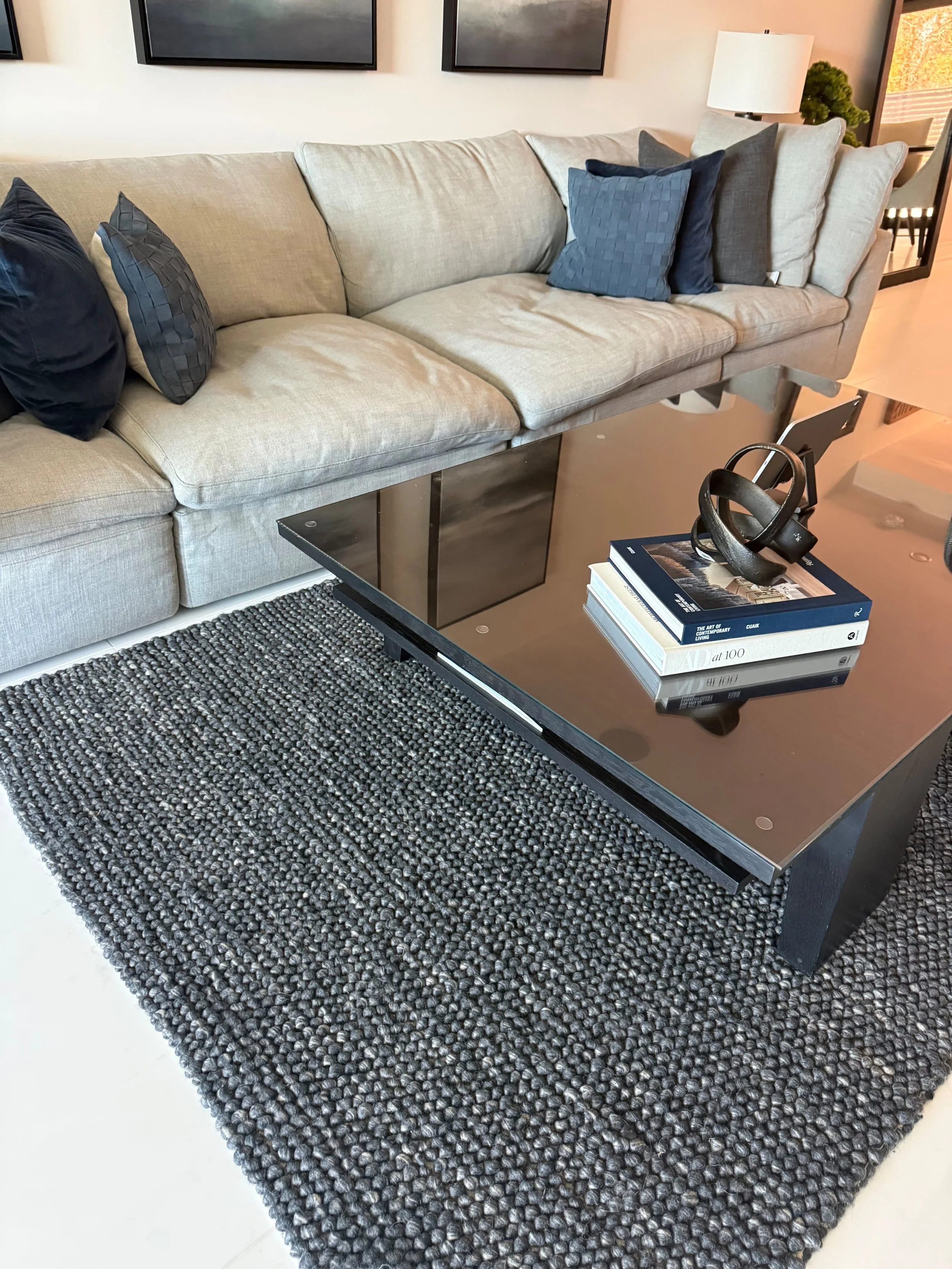Living room with a beige sofa decorated with dark blue and gray pillows, a black coffee table with books and headphones, a black and gray textured rug, a white lamp, and black wall art.