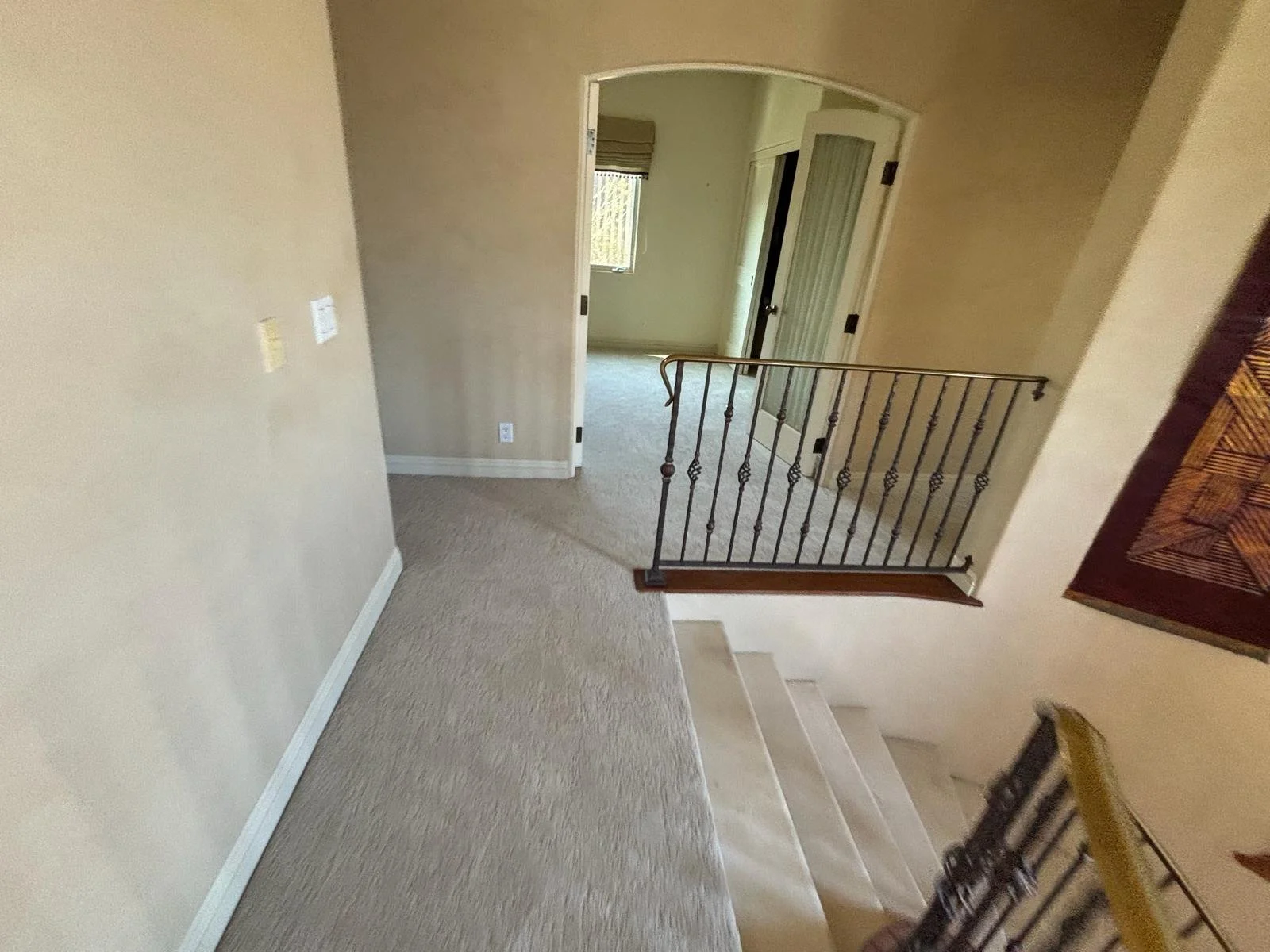 Interior view of a staircase landing with beige carpet, a staircase with wooden steps, a decorative black metal railing, and doorways leading to rooms with windows, beige walls, and window blinds.