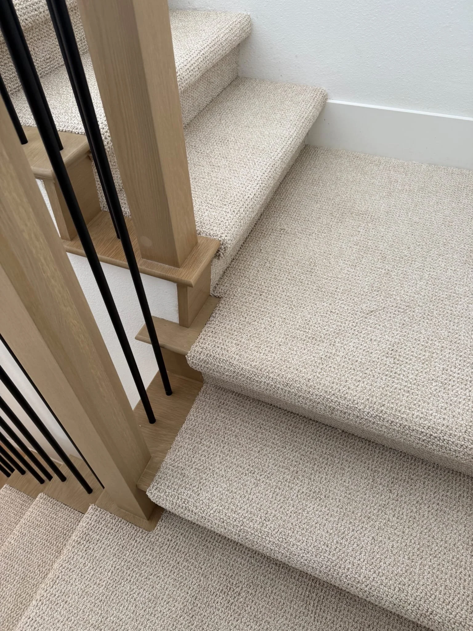 Close-up view of carpeted stairs with beige carpeting, wooden handrail, and black metal balusters.
