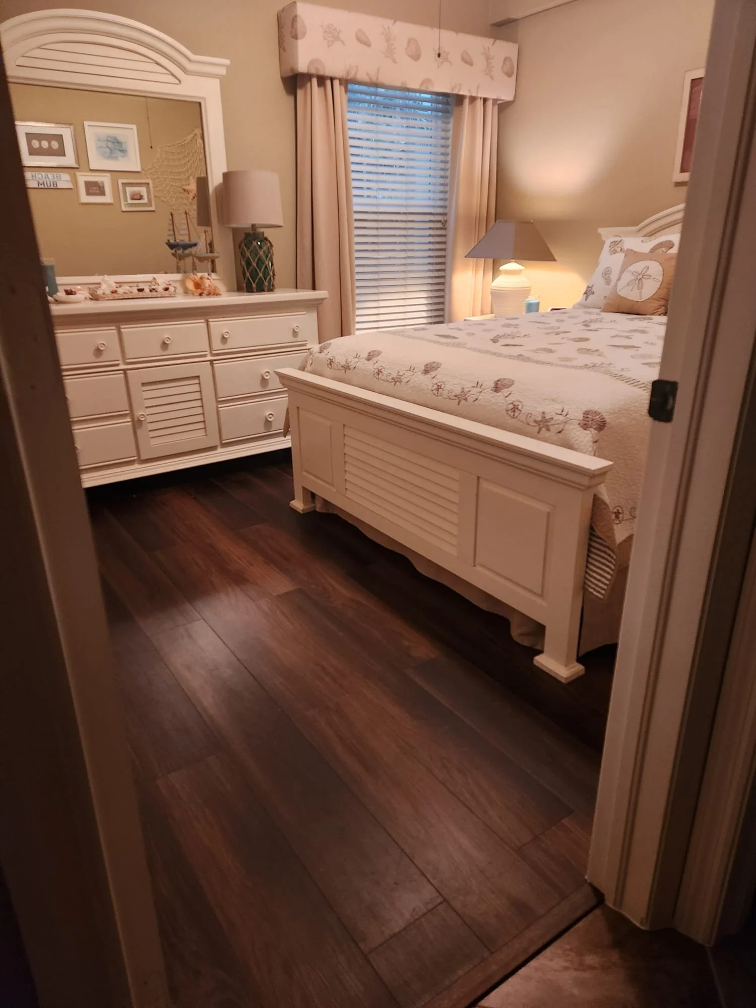 A cozy bedroom with a white wooden bed, dresser with a mirror, and decorative items. The room has beige walls, beige curtains, and a wooden floor. A window with blinds is visible behind the bed.