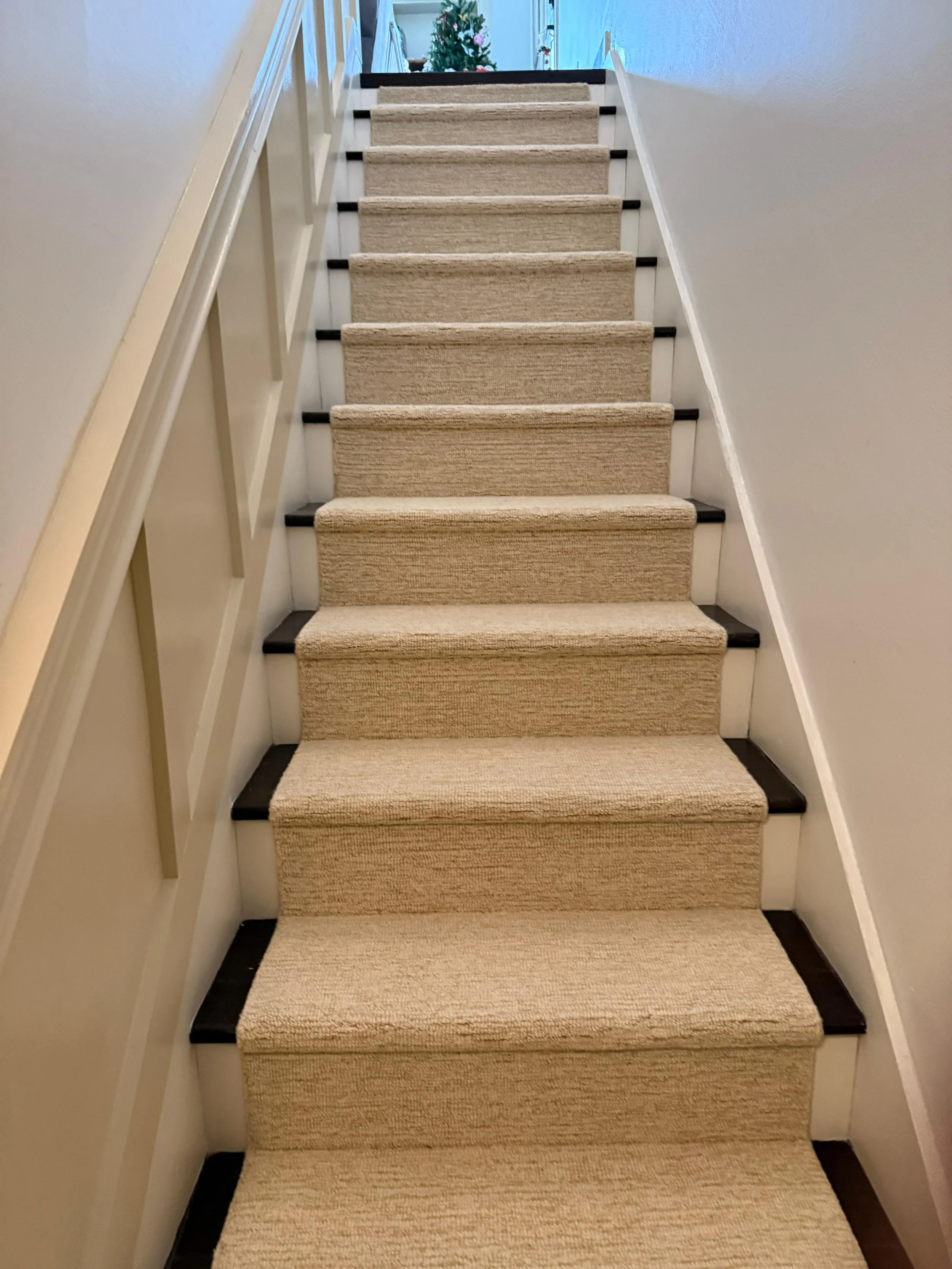 Carpeted staircase with beige carpet and white-painted walls on both sides, leading up to a landing with a Christmas tree decorated with pink ornaments in the background.