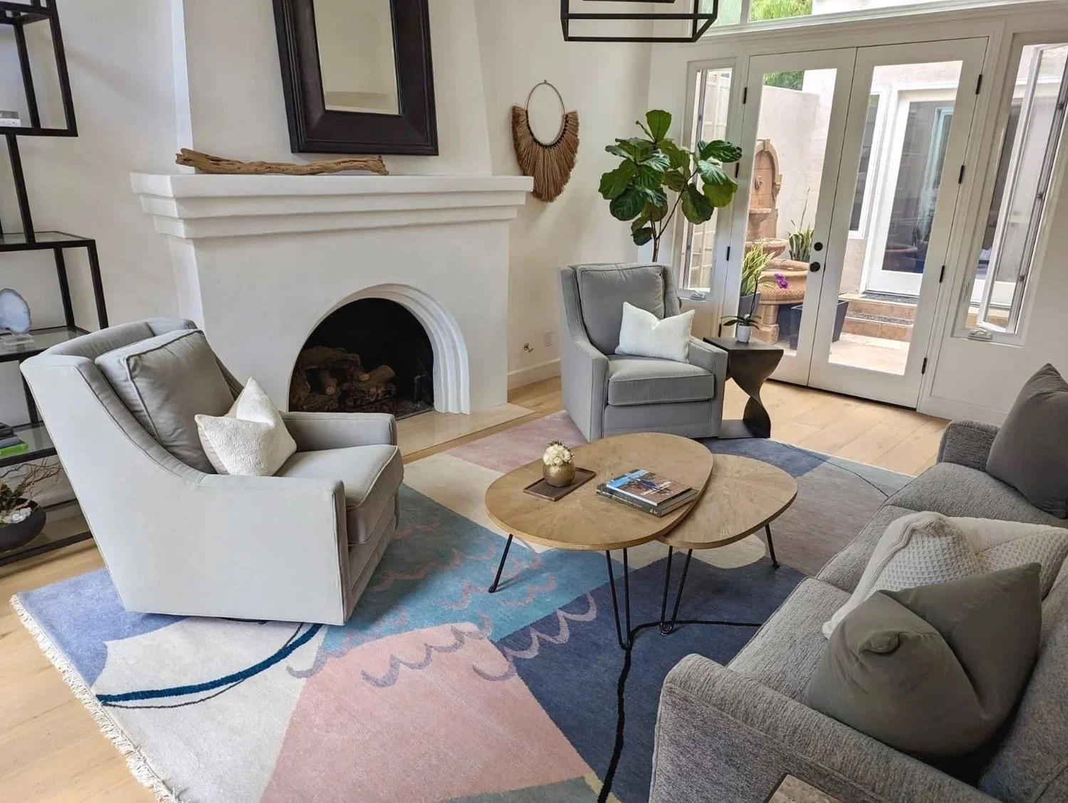 Living room with white fireplace, gray armchairs, a beige sofa, a wooden coffee table, a colorful rug, potted plants, and glass double doors leading to an outdoor patio.