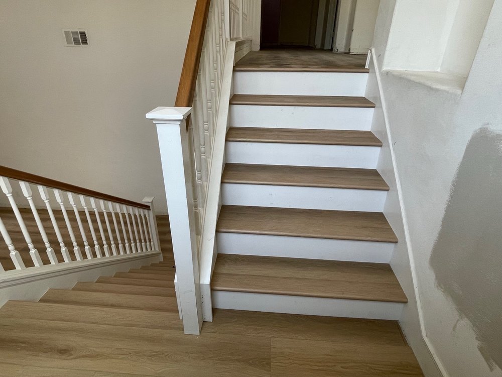 Interior view of a staircase with white risers, wooden treads, and a wooden handrail, leading to an upper floor in a house.