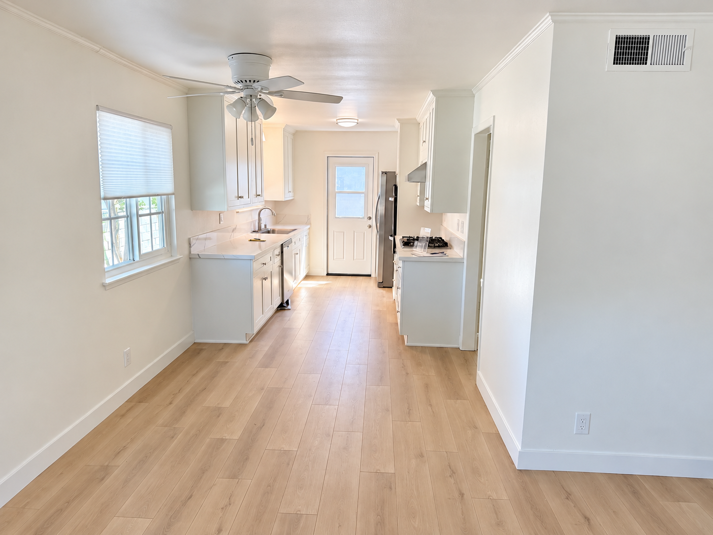 A bright, empty kitchen with white cabinets, a ceiling fan, light wood flooring, a window with blinds, a door, and stainless steel appliances.
