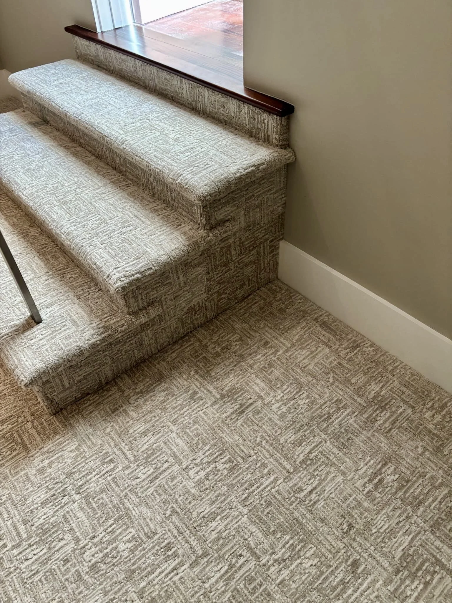 Close-up of a patterned carpeted staircase with three steps, adjacent to a beige wall with white baseboard, and a windowsill with a wooden trim at the top.