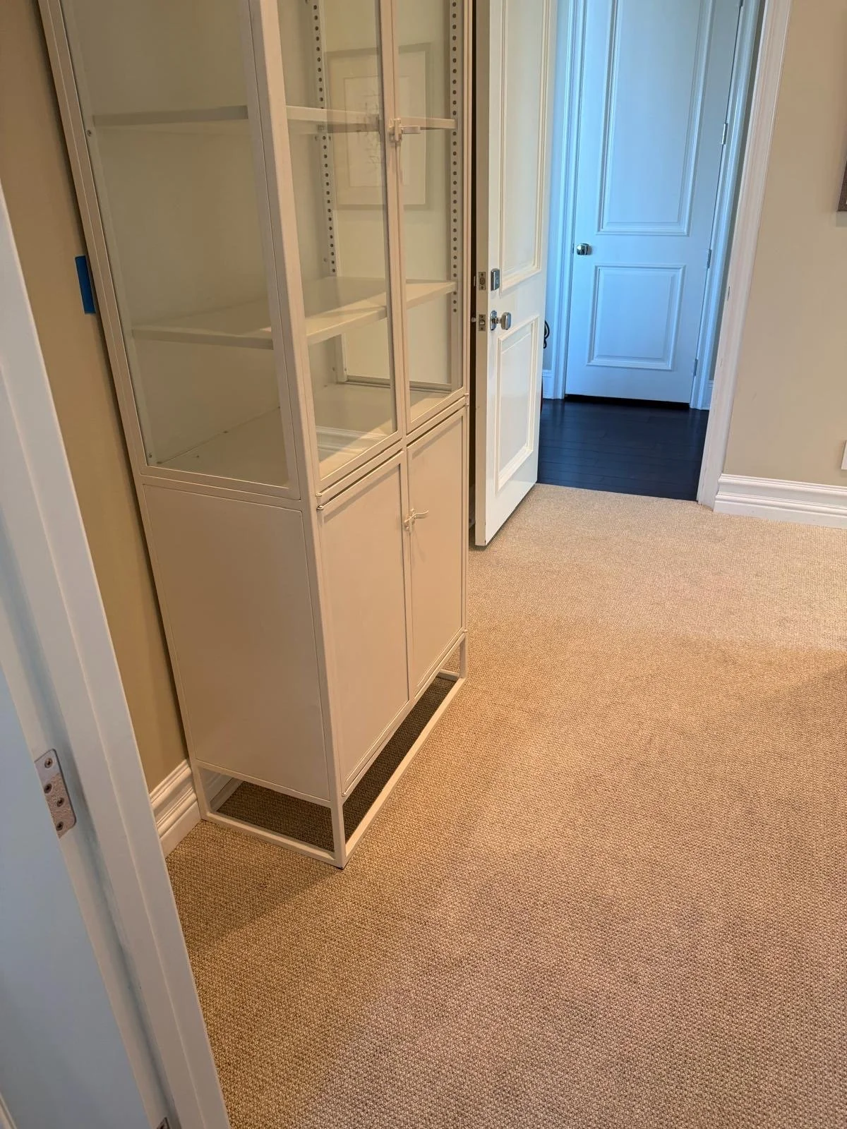 Empty white display cabinet with glass doors in a room with beige carpet, open door with dark wood flooring visible beyond.