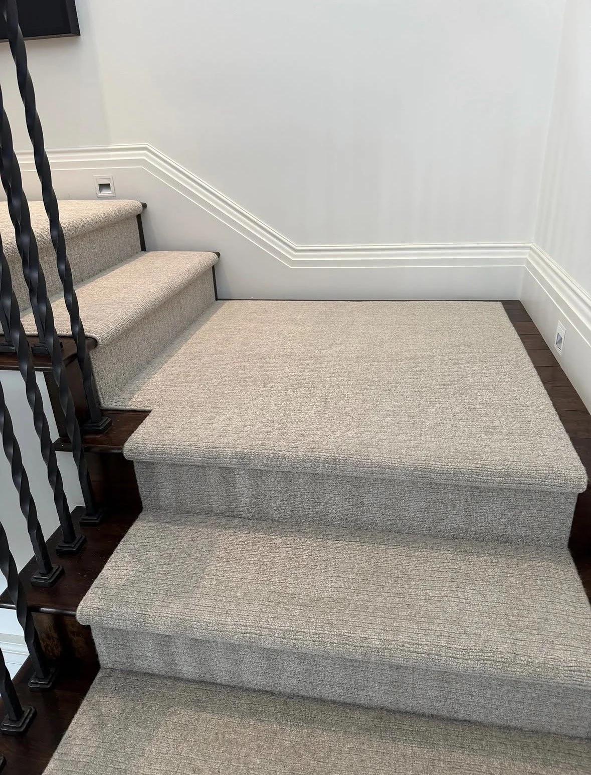 Carpeted staircase with beige carpet, black metal railing, white walls, and wall outlet, leading to a higher level in a building.