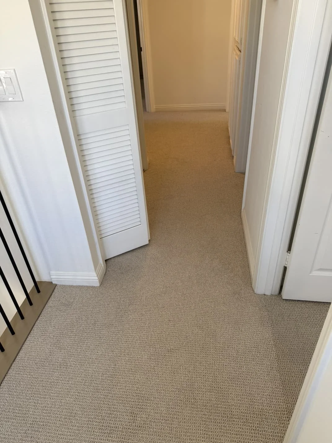Photo of a hallway with beige carpet, white walls, and multiple doors, including a louvered closet door and open doorway leading into other rooms.