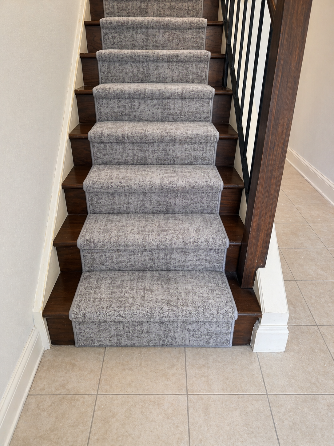 Wooden staircase with a grey carpet runner, beige tiled floor at the bottom, and a black metal railing on the right side.
