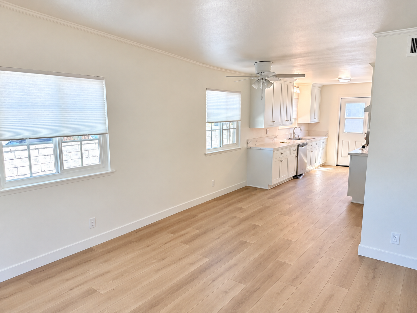 Empty living area with light wood flooring, white walls, windows with blinds, and a white kitchen with cabinets and a ceiling fan.