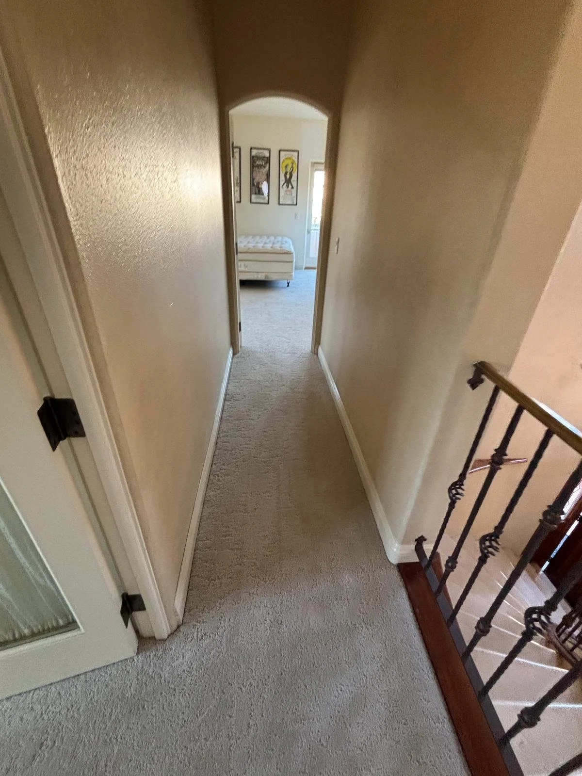 A hallway with beige walls, carpeted floor, and a staircase leading downstairs, with a bedroom visible at the end of the hallway, decorated with framed artwork on the wall.