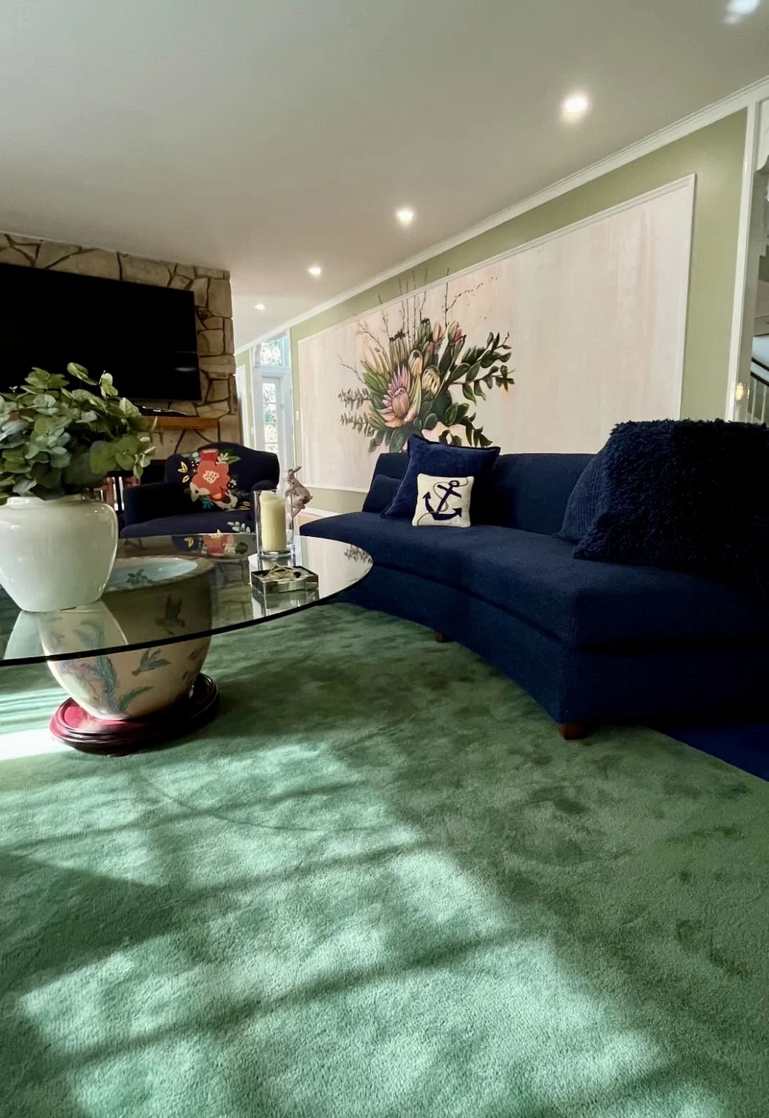 Living room with a green carpet, navy blue couches, a glass coffee table, and a large floral painting on the wall.
