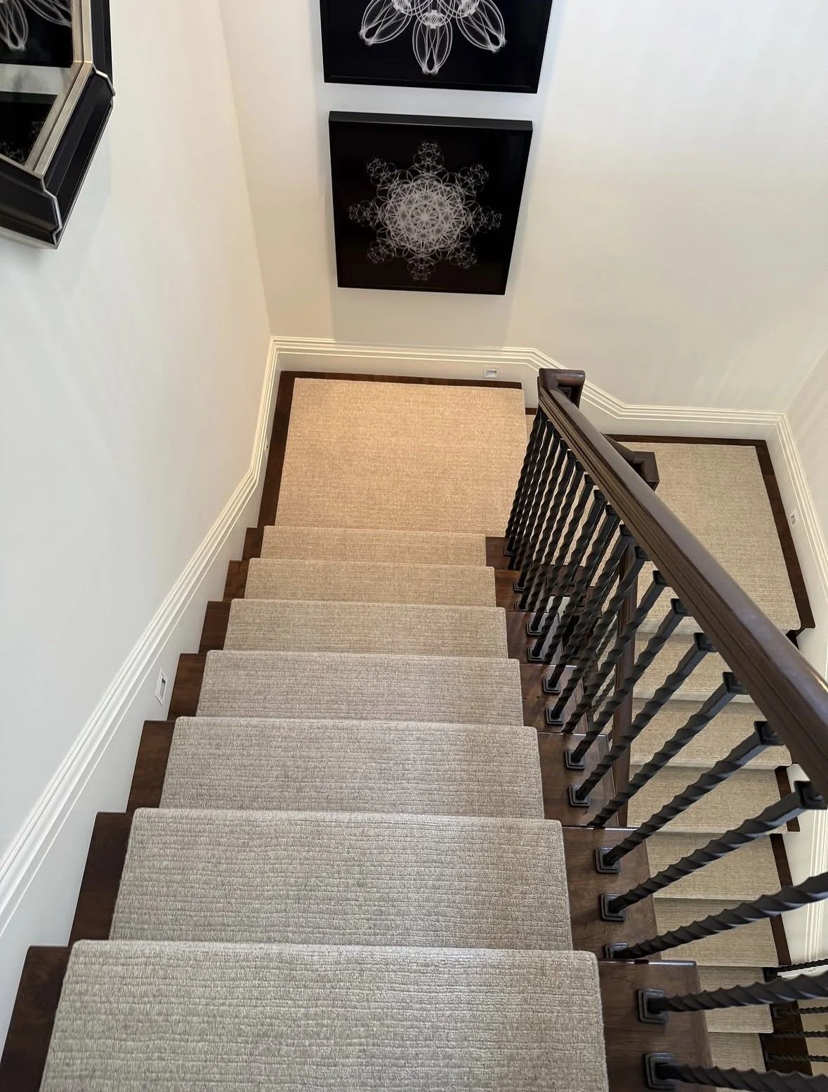 View from the top of a staircase with beige carpeted steps, a dark wood handrail, and black balusters. At the bottom, there are two framed black and white geometric art pieces on the wall.