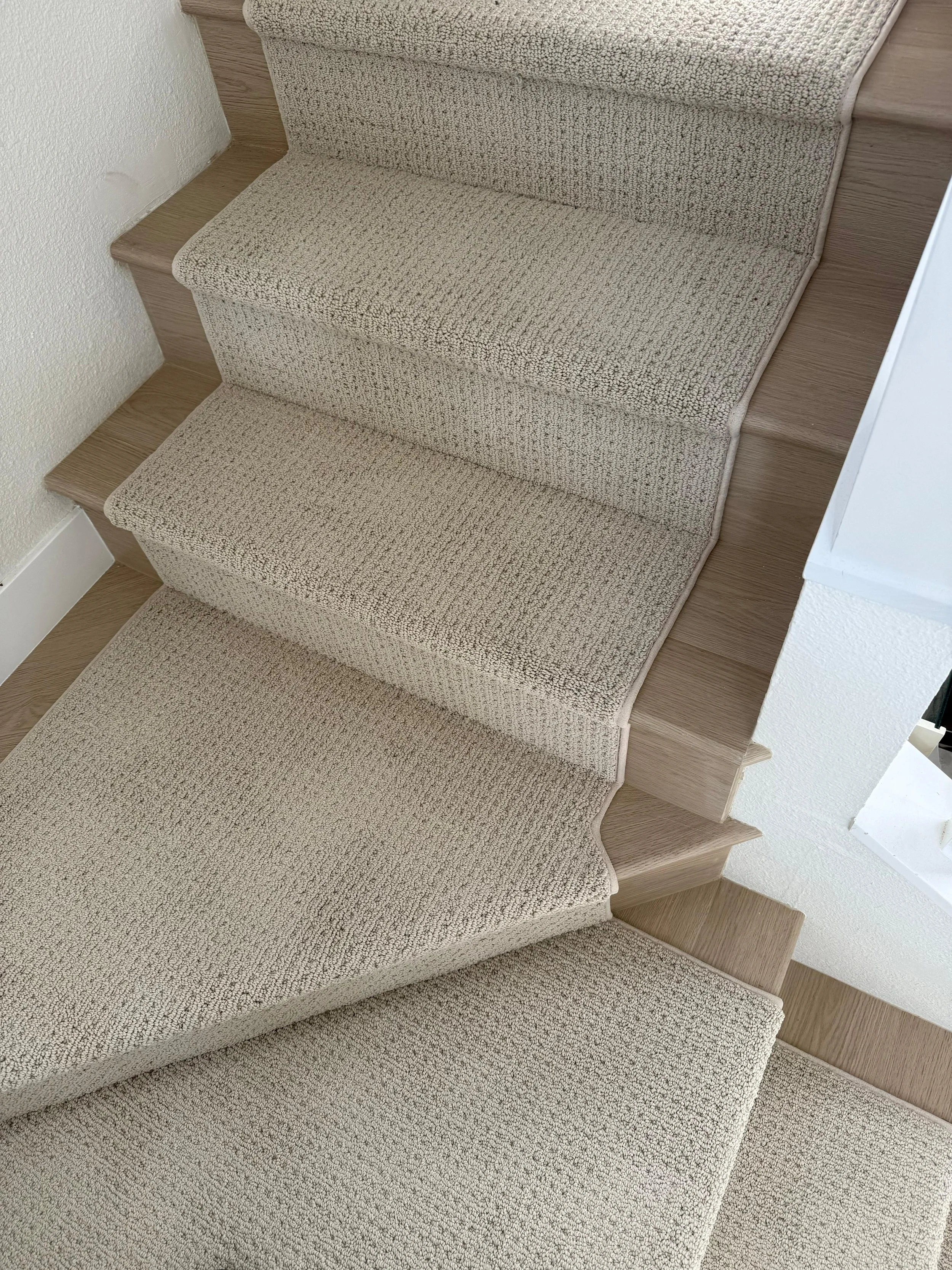 Carpeted staircase with beige carpet on steps and light wood risers, next to white textured wall and a white window ledge.