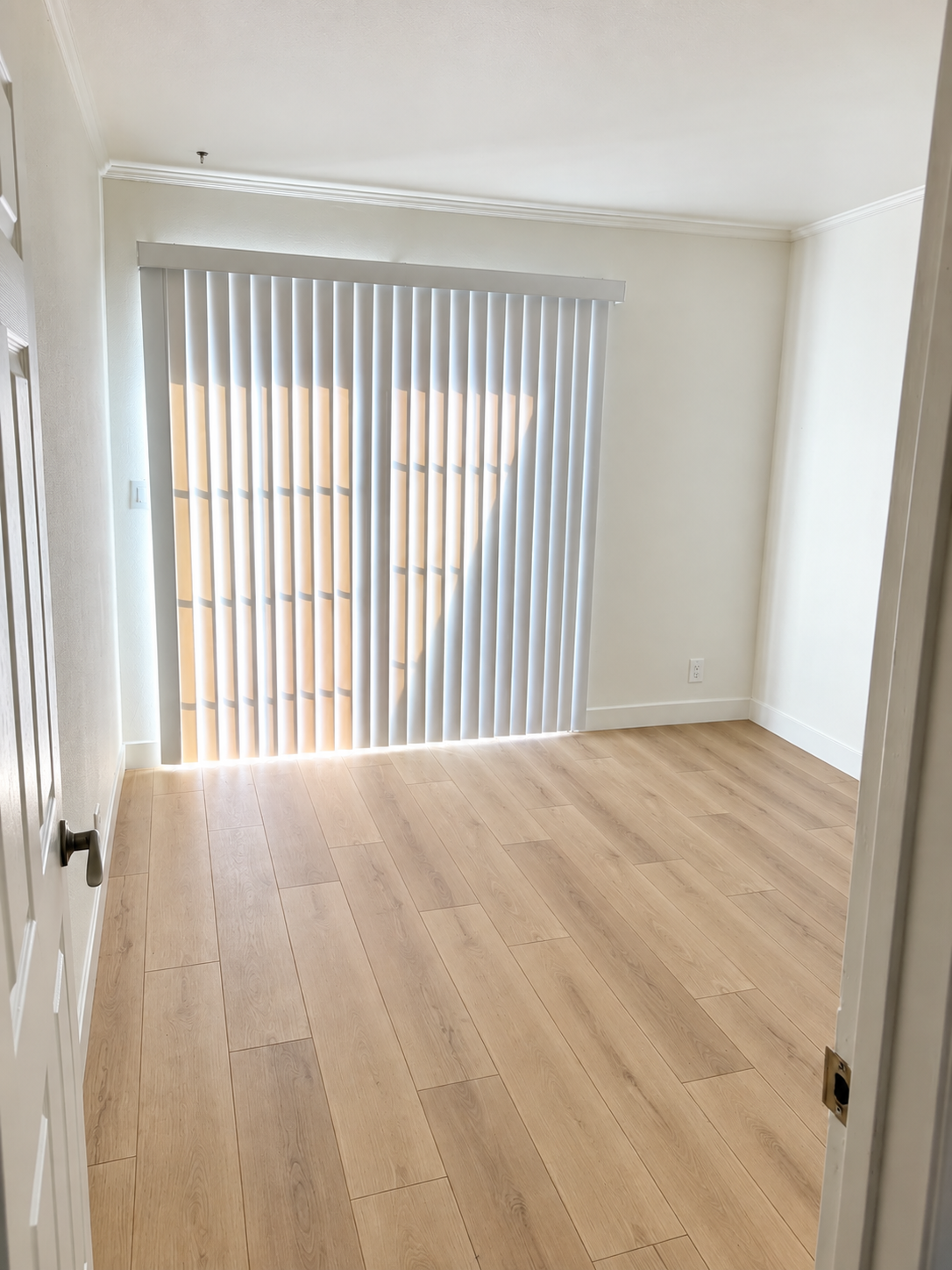Empty room with natural wood flooring, white walls, and vertical blinds covering a window with sunlight streaming through.