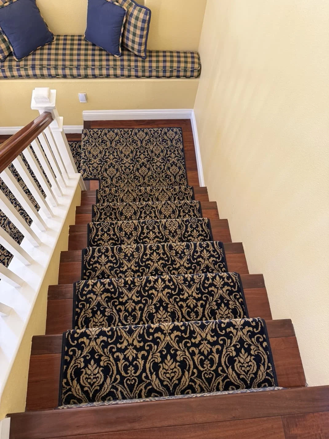 A staircase with dark ornate patterned carpet runner, wooden steps, yellow walls, and a built-in window seat with checkered cushion and two blue pillows.