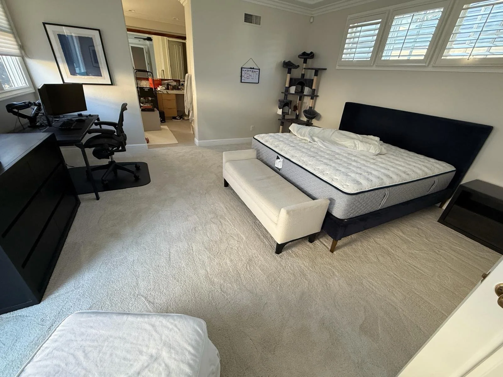 A bedroom with a bed frame and mattress, a bench at the foot of the bed, and a cat tree near the window with blinds. Also, a computer desk with a chair and a monitor, and a doorway to a bathroom.