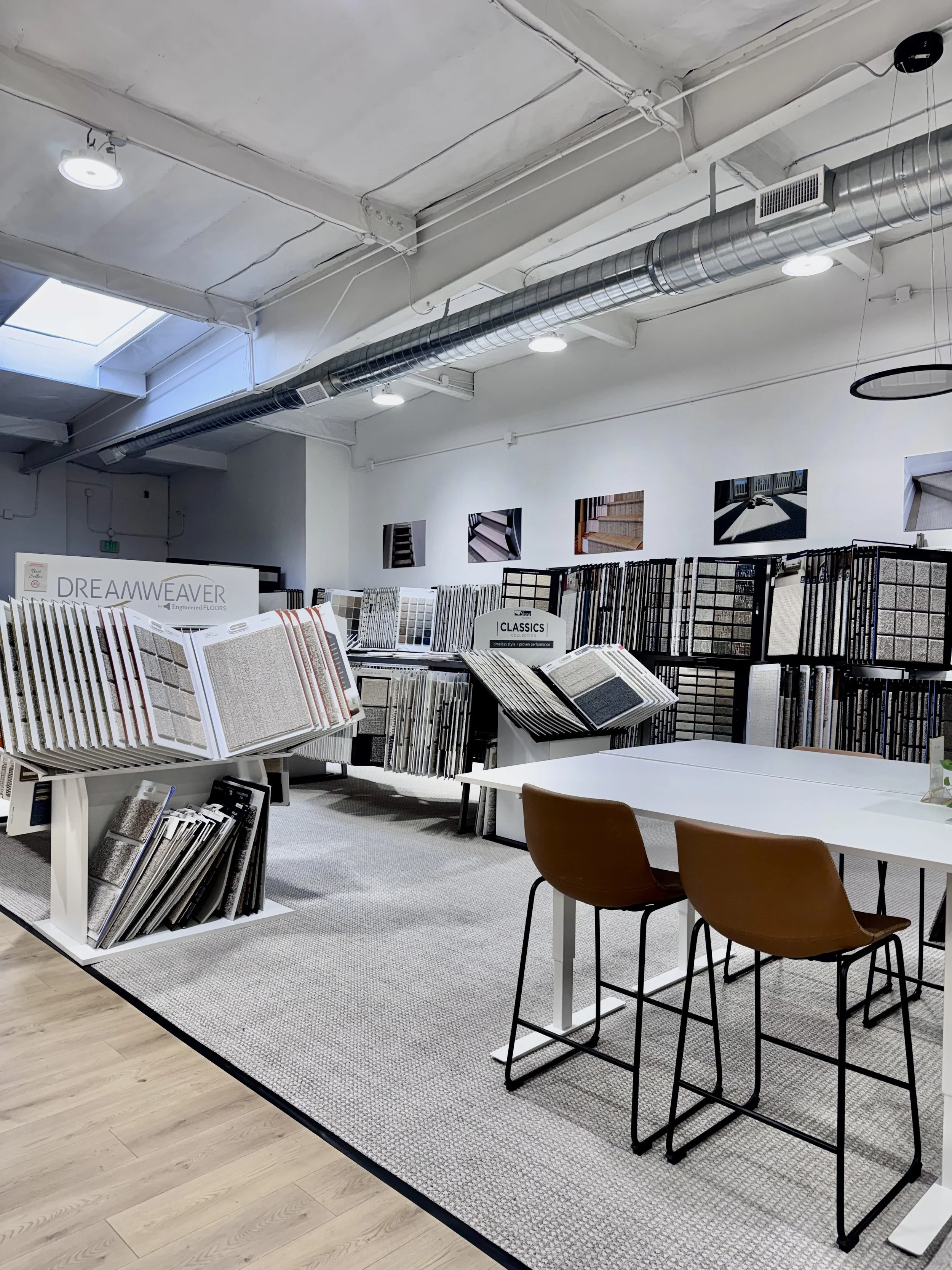 Interior of flooring store with display racks of tile samples, a white table with brown chairs, and framed pictures of flooring on the wall.