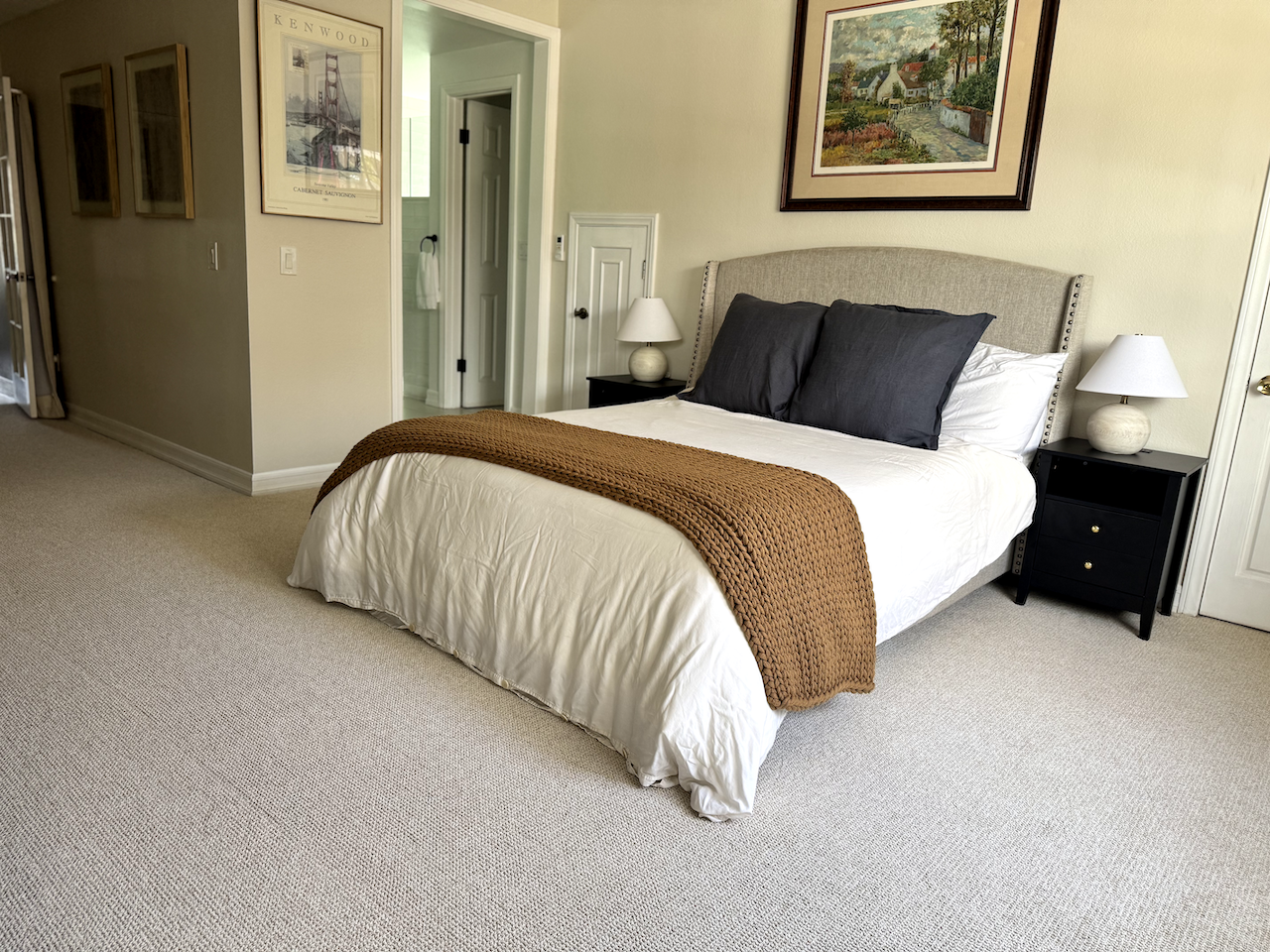 A neatly made bed with a beige upholstered headboard, white bedding, two dark gray pillows, and a brown knitted throw blanket. There are two black nightstands with white lamps on each side of the bed. A framed painting of a scenic country road hangs 