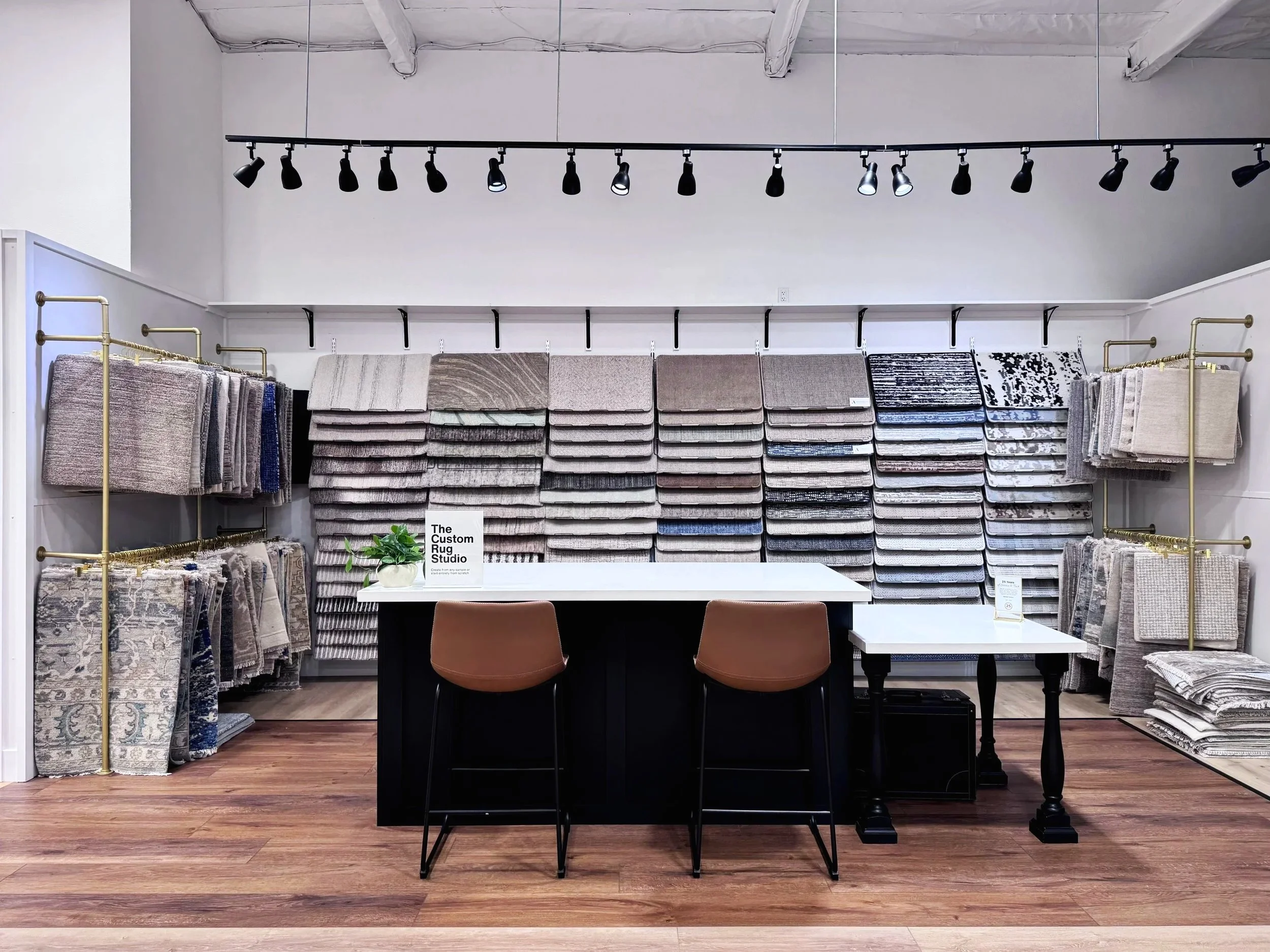 Display of numerous folded rugs and carpets on shelves and racks in a showroom, with a table and chairs in front, and spotlights overhead illuminating the space.