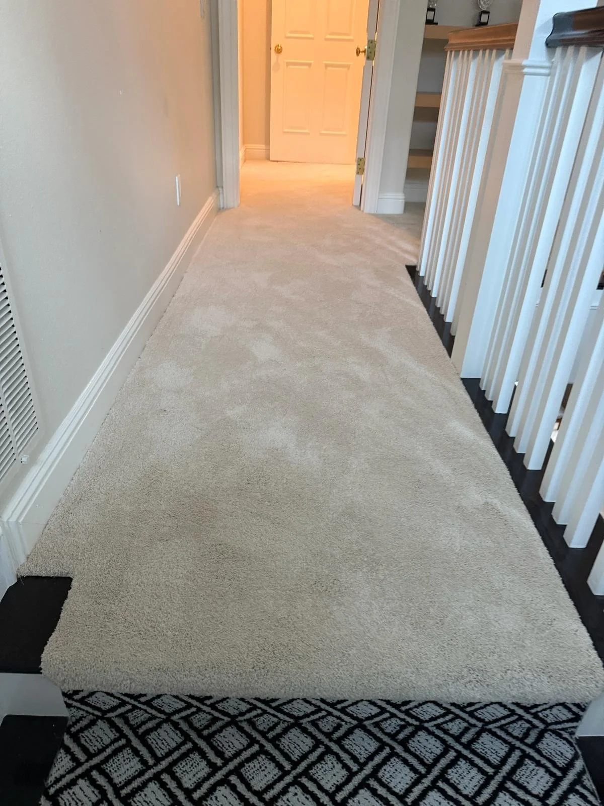 Hallway with beige carpet runner, white walls, and a staircase with white spindles on the right. There is a door at the end of the hallway, slightly ajar, with a room visible beyond.
