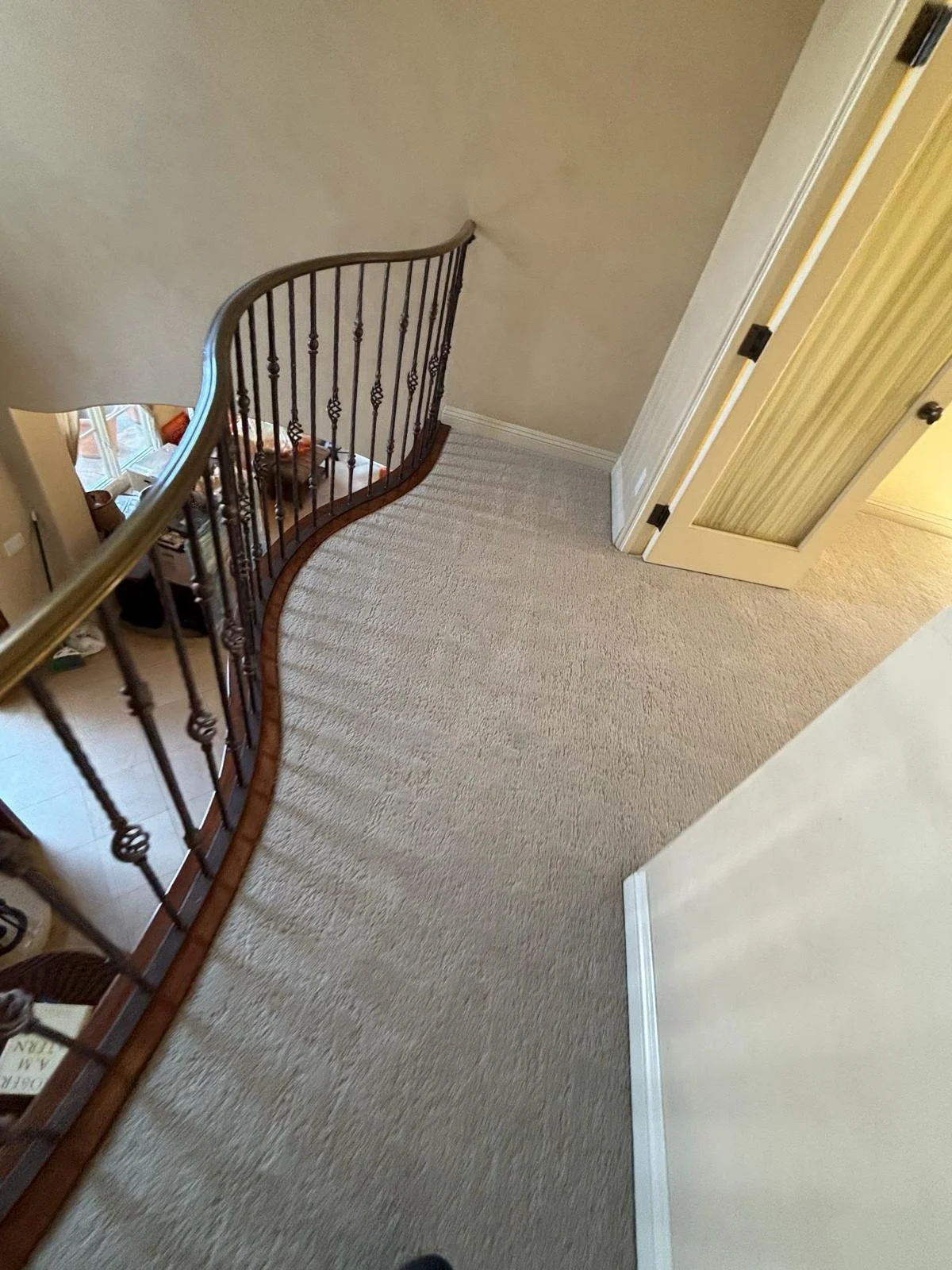 A carpeted upstairs landing with a curved wooden and metal railing, beige walls, and an open door leading to a room with a large window.