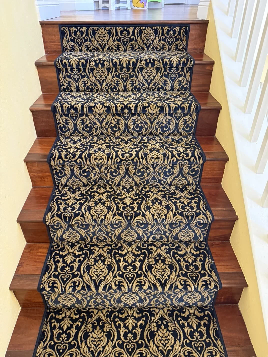 Staircase with a black and gold ornate patterned carpet runner, wooden steps, yellow walls, and a white railing on the right.