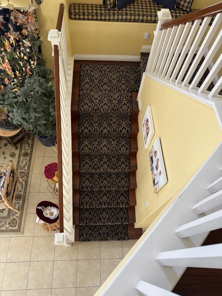 View of a staircase from the top, showing a decorative black and gold patterned runner rug, white railings, and a yellow wall with framed photos. To the left, part of a floral plant, a small wooden table with a red cloth, and tan tiled floor. Part of