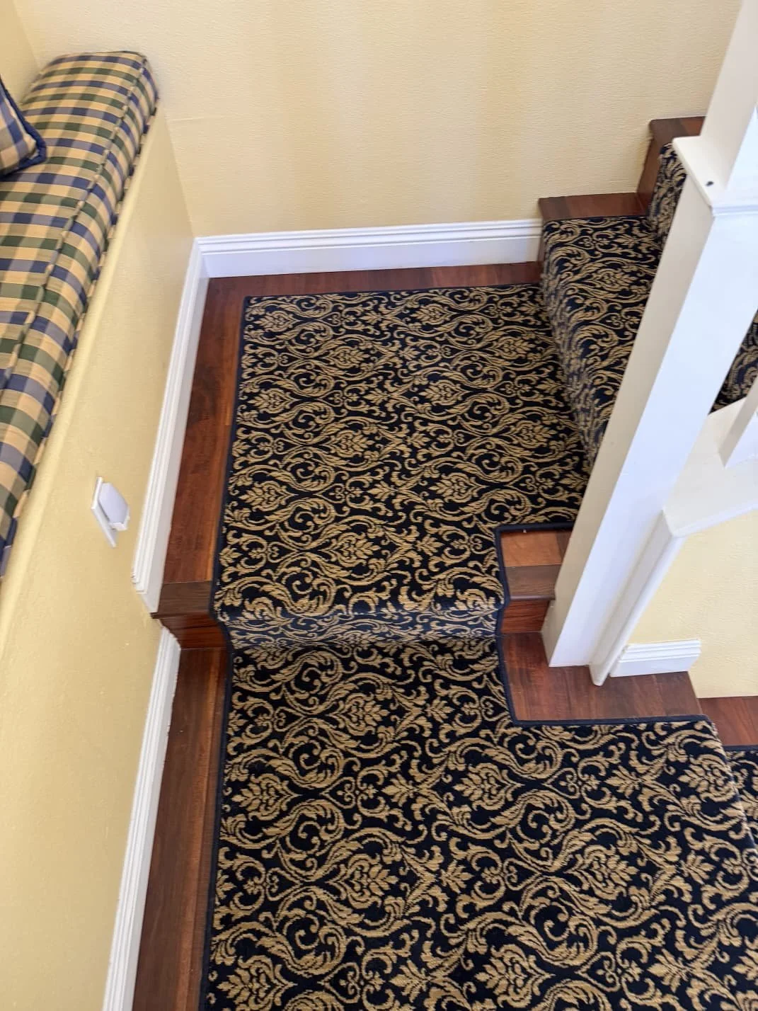 A stair carpet runner with an ornate black and gold floral pattern on a staircase, adjacent to a yellow wall with a checked cushioned seat on the left and white baseboards.