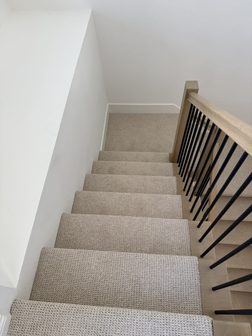 Top view of a staircase with beige carpeted steps and a wooden handrail with black balusters, against white walls.
