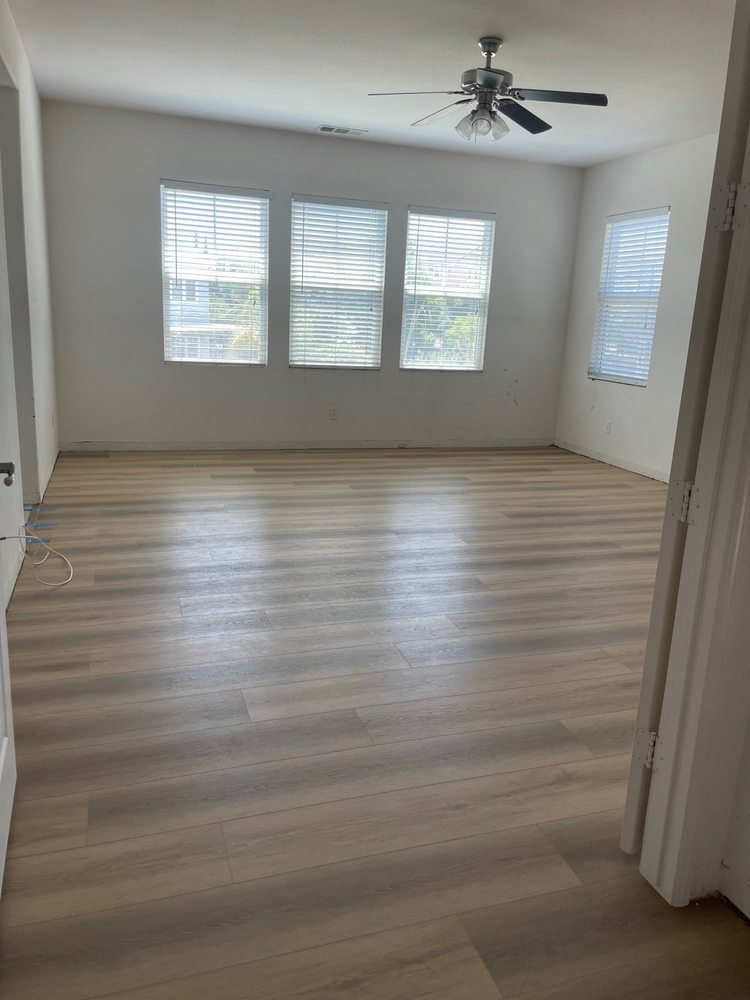 Empty room with light-colored wood flooring, white walls, four windows with blinds, ceiling fan, and partially visible door.