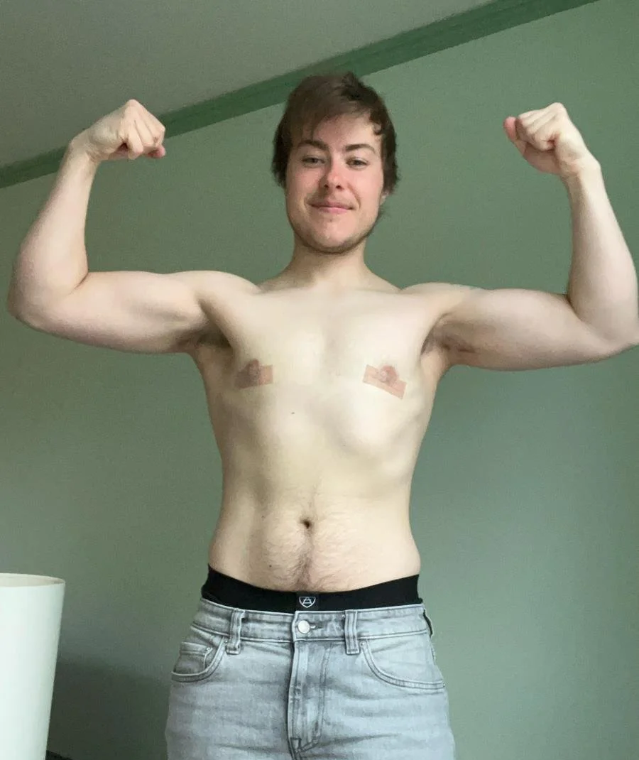 A shirtless young man with brown hair flexing his biceps and smiling at the camera, standing against a light green wall.