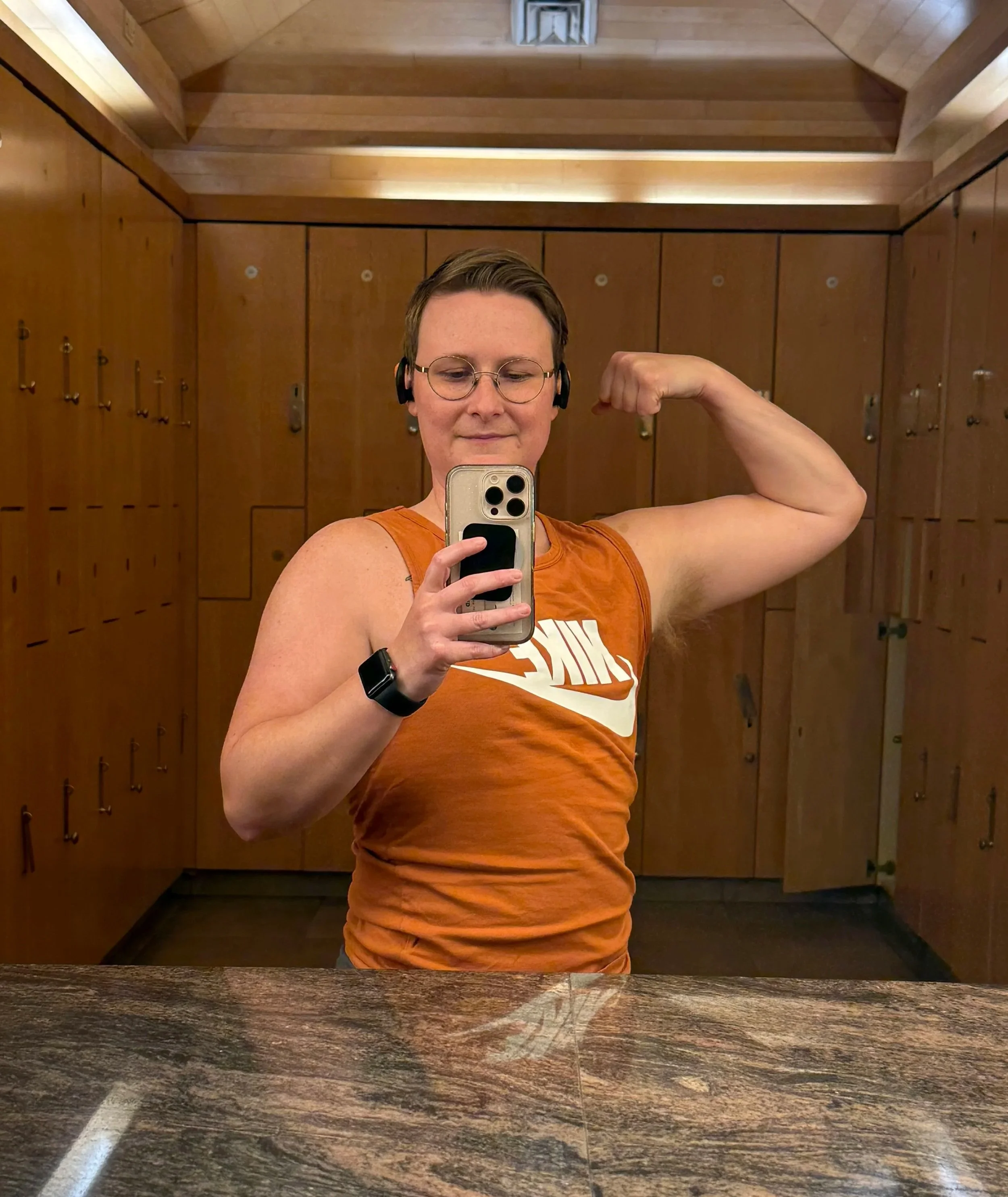 A woman taking a mirror selfie in a wooden locker room, flexing her right arm, wearing glasses, headphones, and an orange sleeveless Nike shirt.
