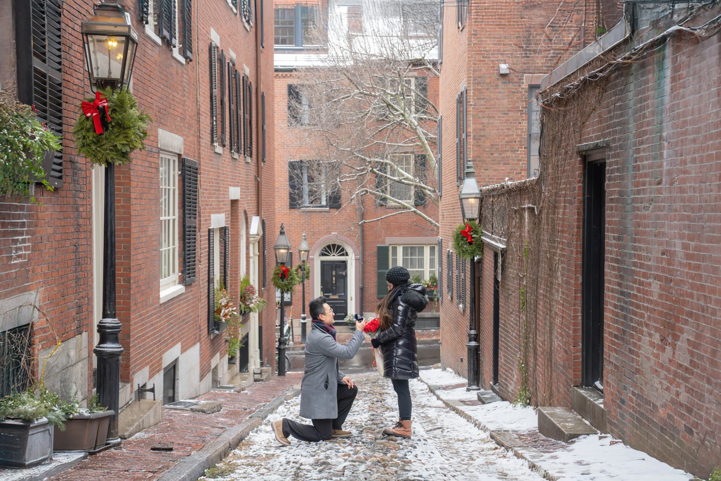 A couple on a snow-covered brick alleyway surrounded by red brick buildings, sharing a moment of celebration or proposal, with festive wreaths and lights adorning the scene.
