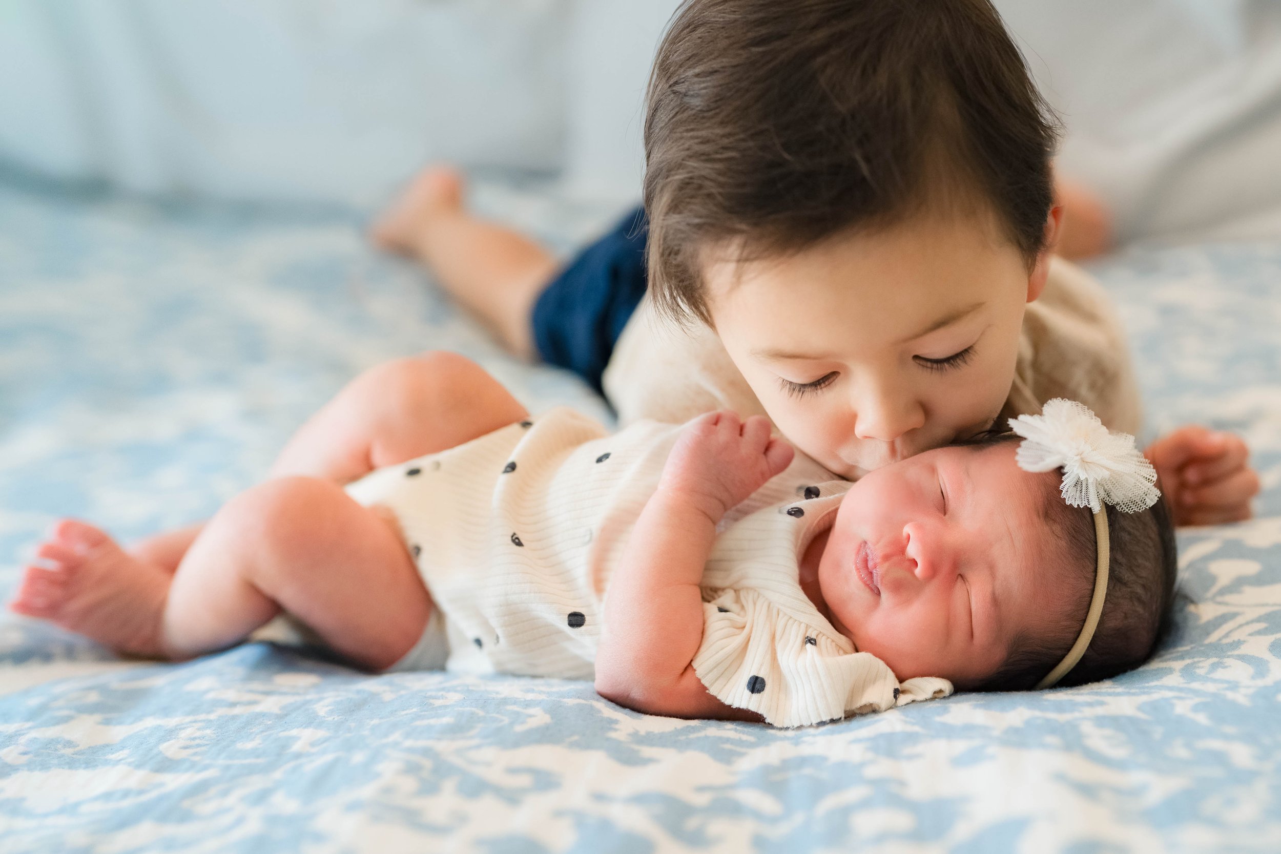 A young boy and a newborn baby lying on a blue and white patterned blanket, with the boy gently kissing the baby's forehead.