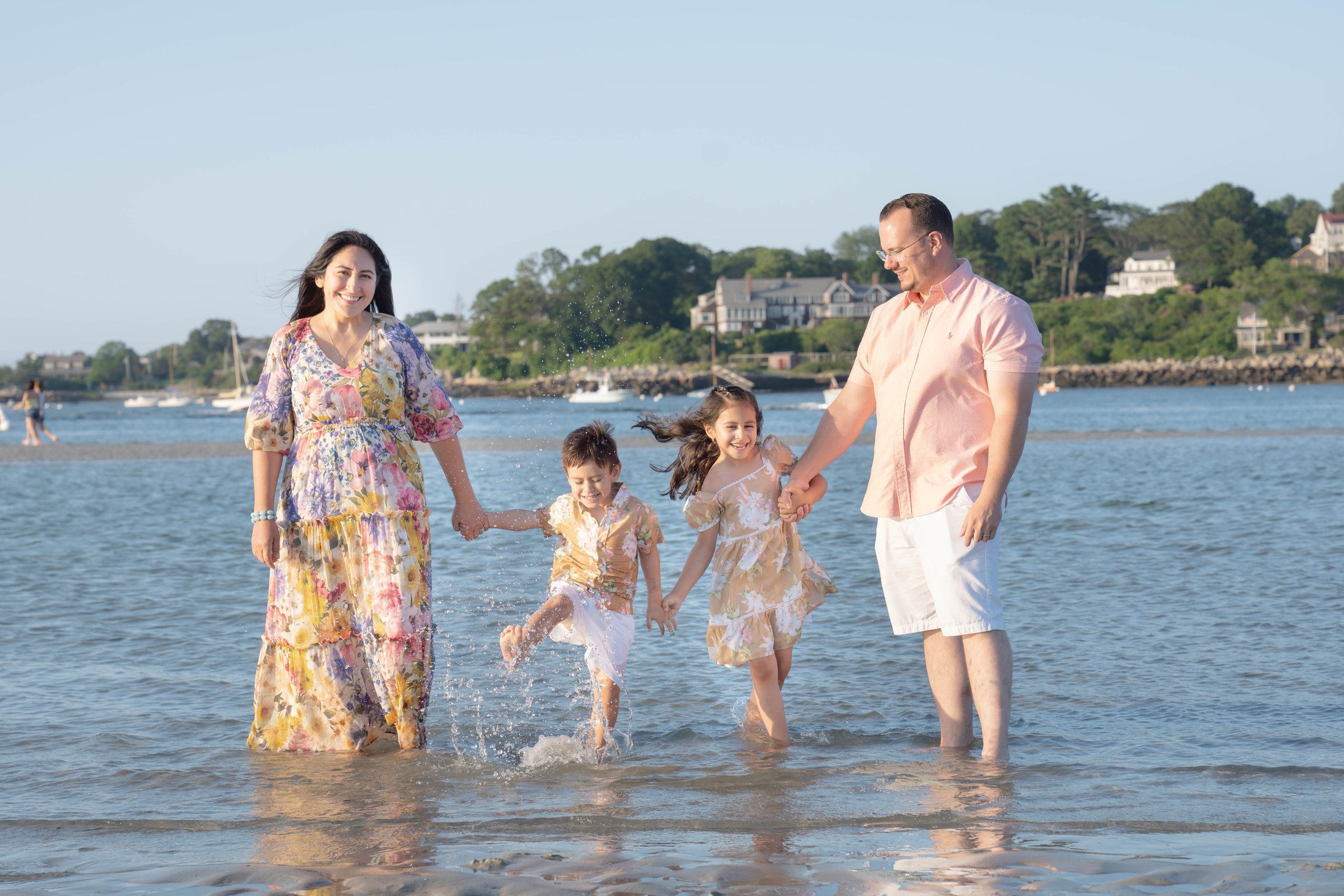 A family of five enjoying a day at the beach, holding hands and playing in the water with houses and trees in the background.