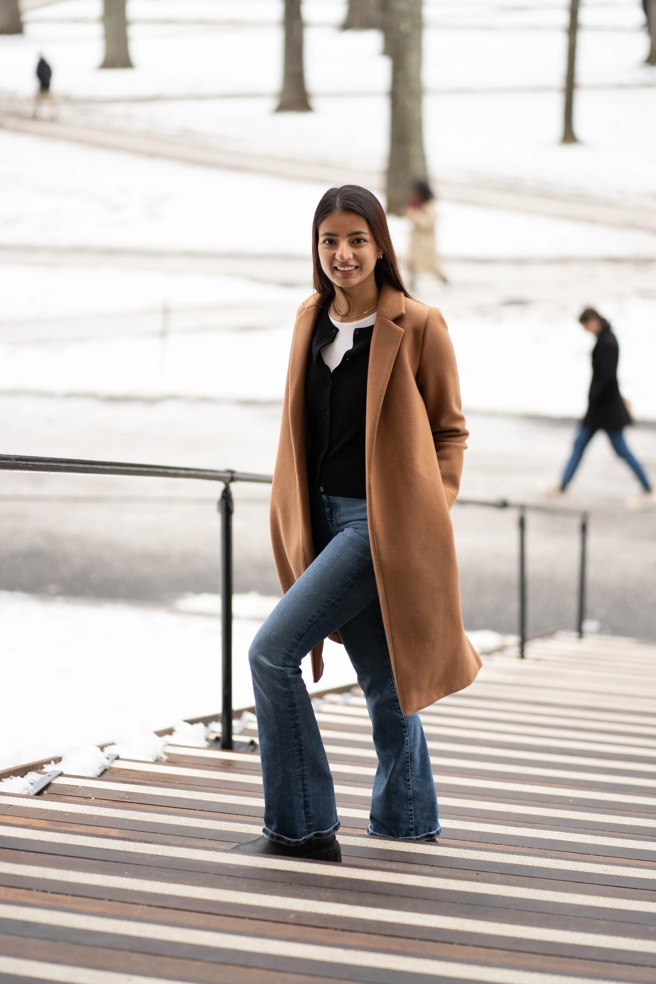 Young woman standing outdoors on a snowy day with trees in the background, wearing a camel coat, black and white shirt, and jeans, smiling at the camera.
