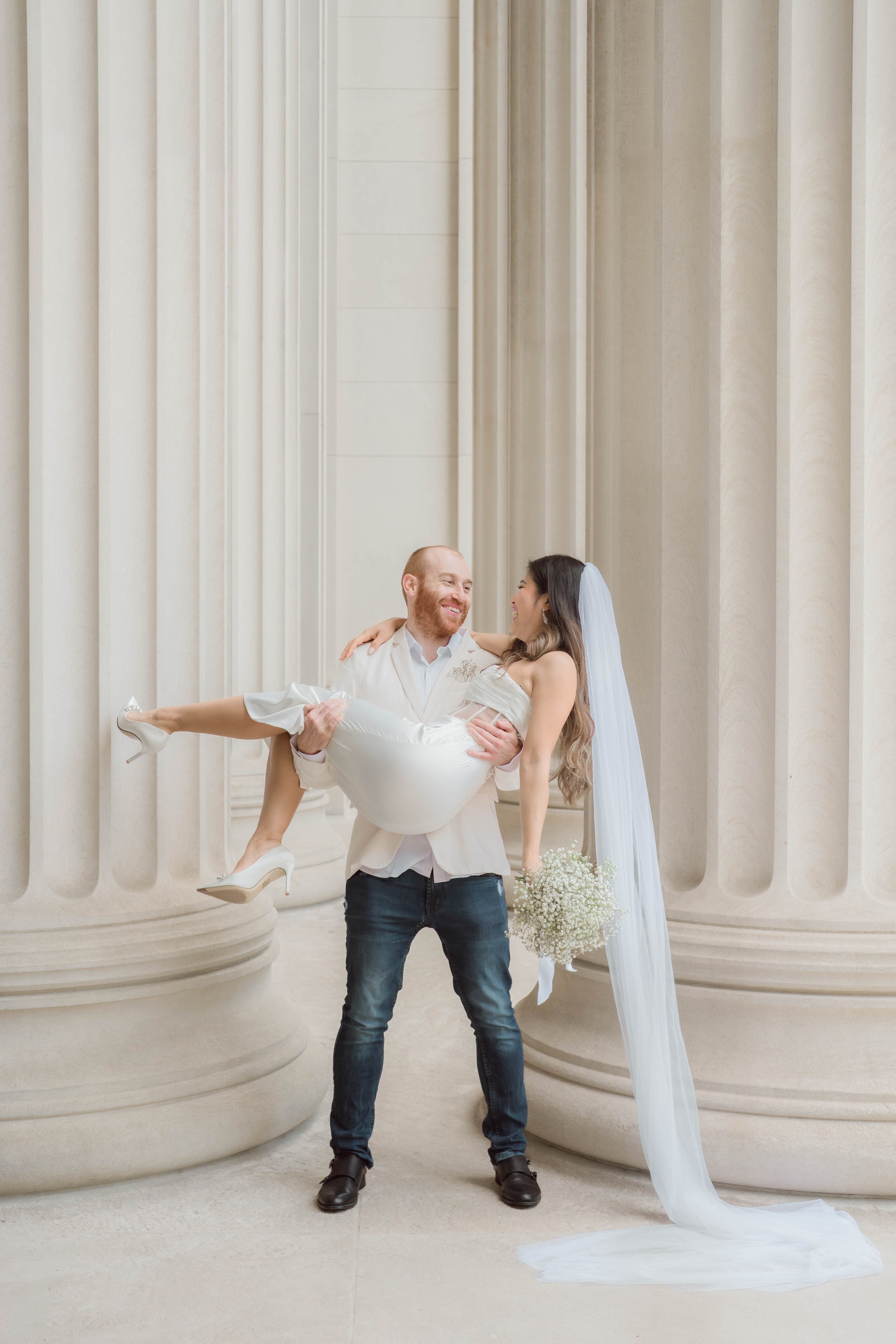 A man holding a woman in a wedding dress and veil in a large, marble architectural setting with tall white columns.