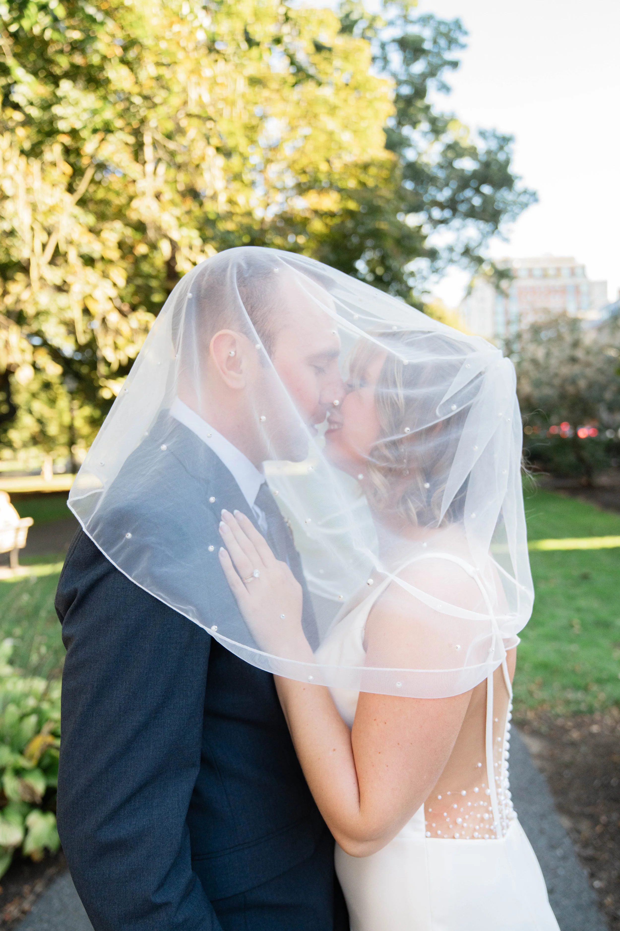 A newlywed couple is sharing a kiss outdoors, with the bride wearing a veil and the groom in a suit. The background features trees and a cityscape.
