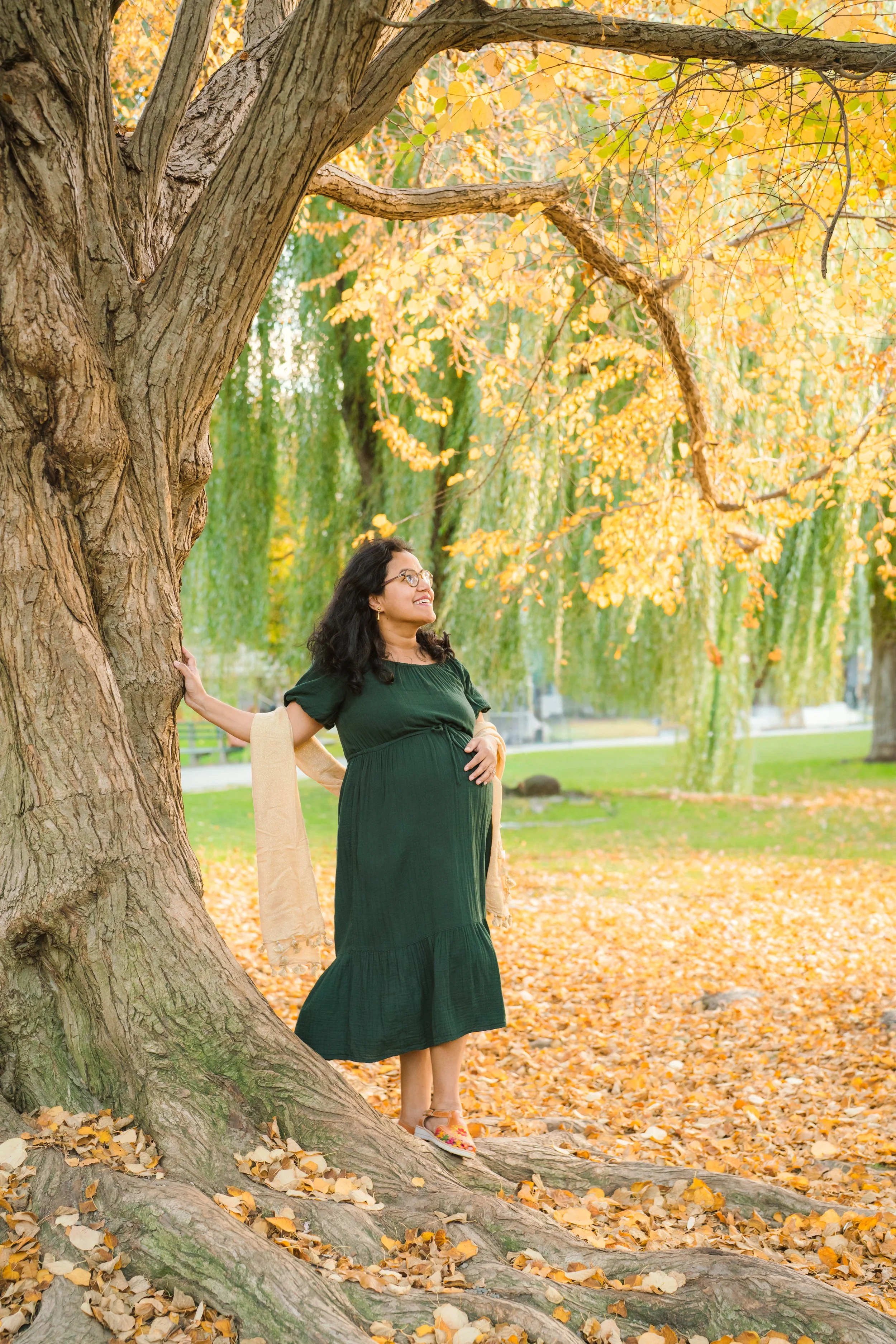 Woman is standing outdoors by a large tree with yellow autumn leaves, wearing a dark green dress and colorful sandals, smiling and touching her pregnant belly.