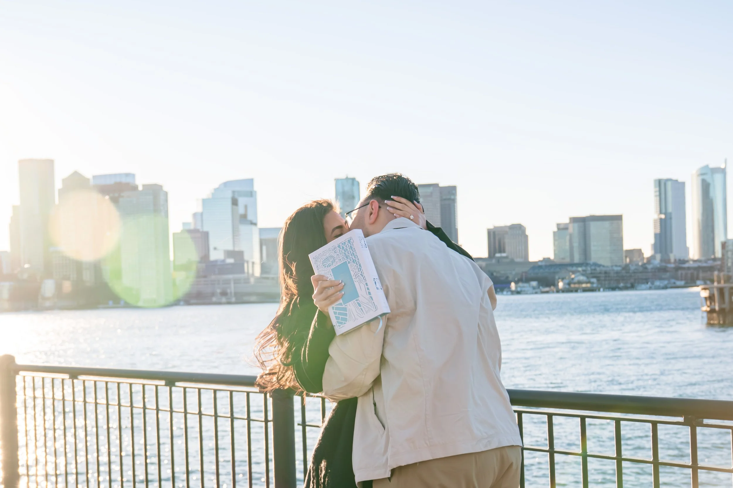 A couple embracing and kissing near a body of water with a city skyline in the background during sunset.