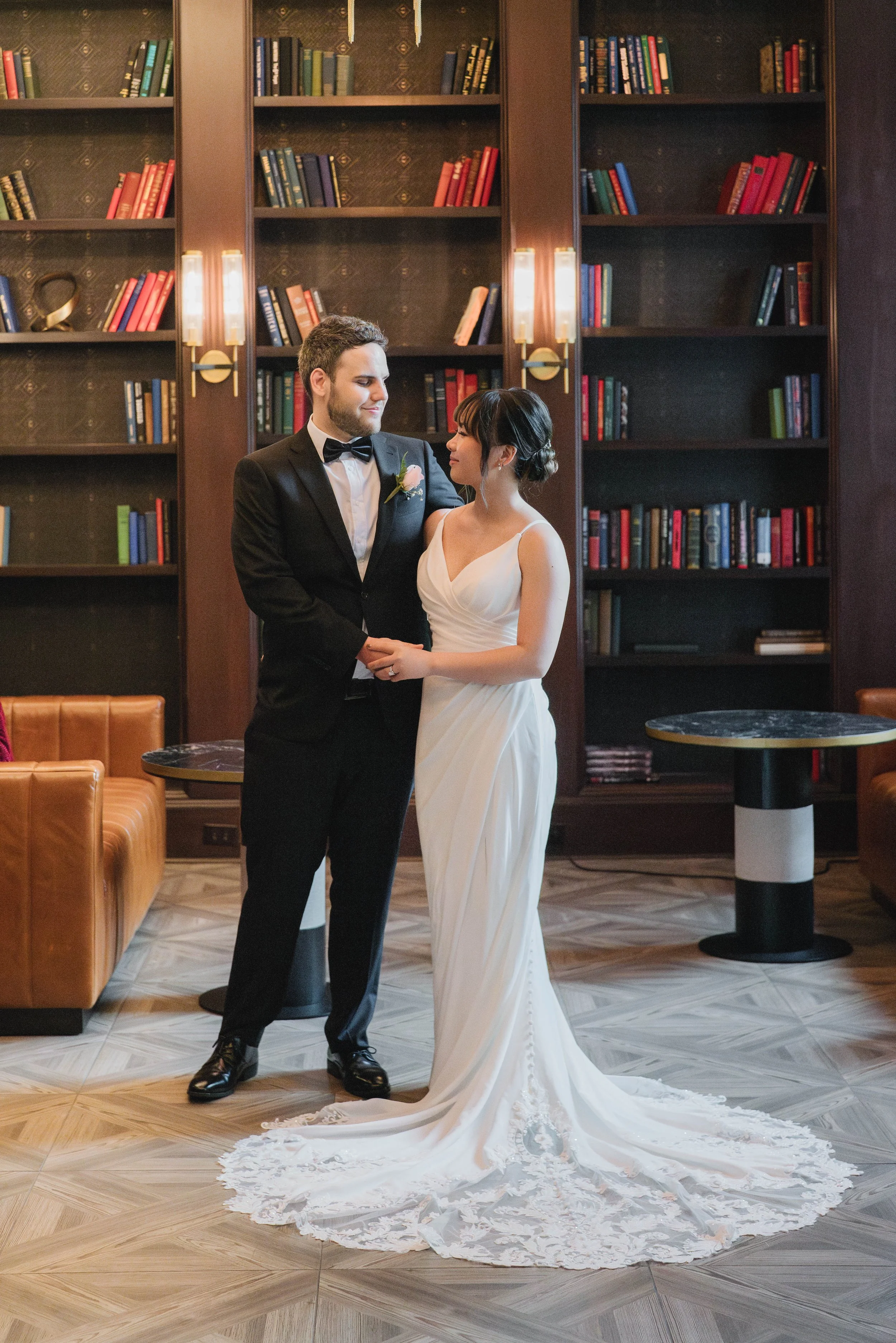 A newlywed couple stands holding hands in an elegant room with dark wooden shelves filled with books. The groom is wearing a black tuxedo with a bow tie and white shirt, and the bride is wearing a white wedding gown with lace details, a train, and he