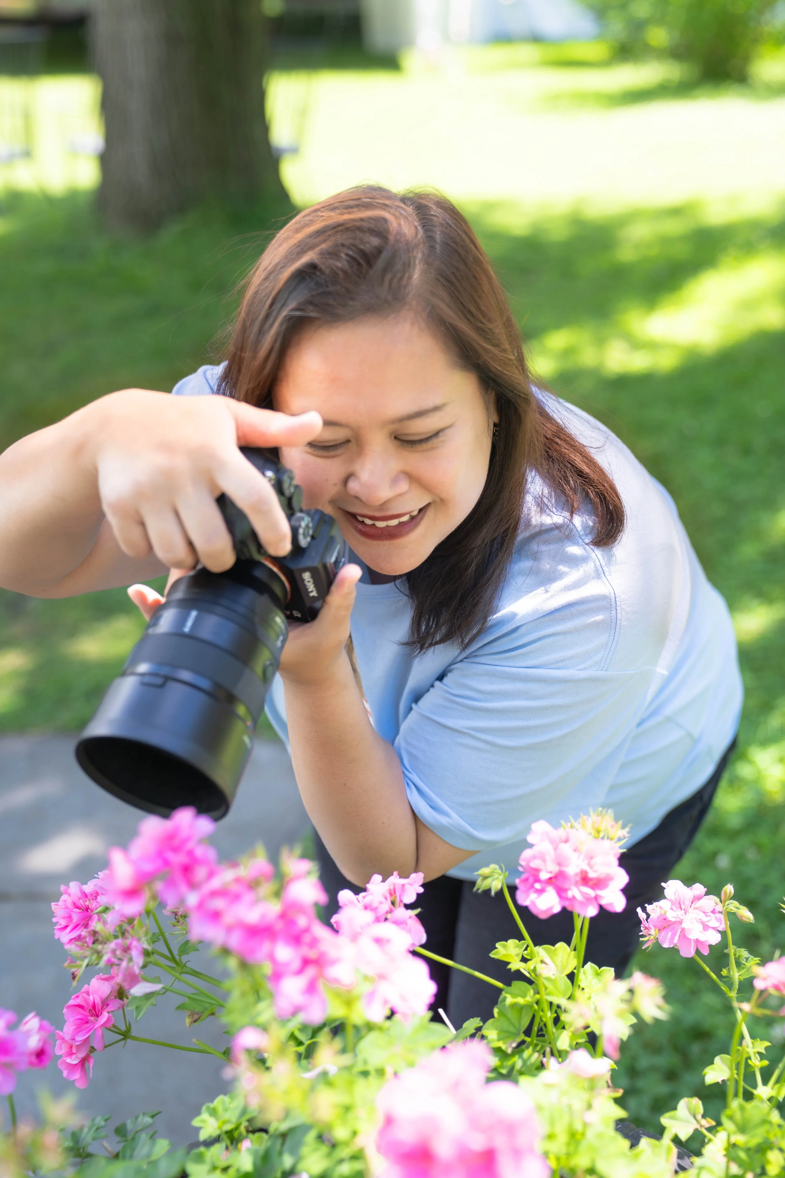 A photo of Mim using a camera to photograph pink flowers outdoors on a sunny day.
