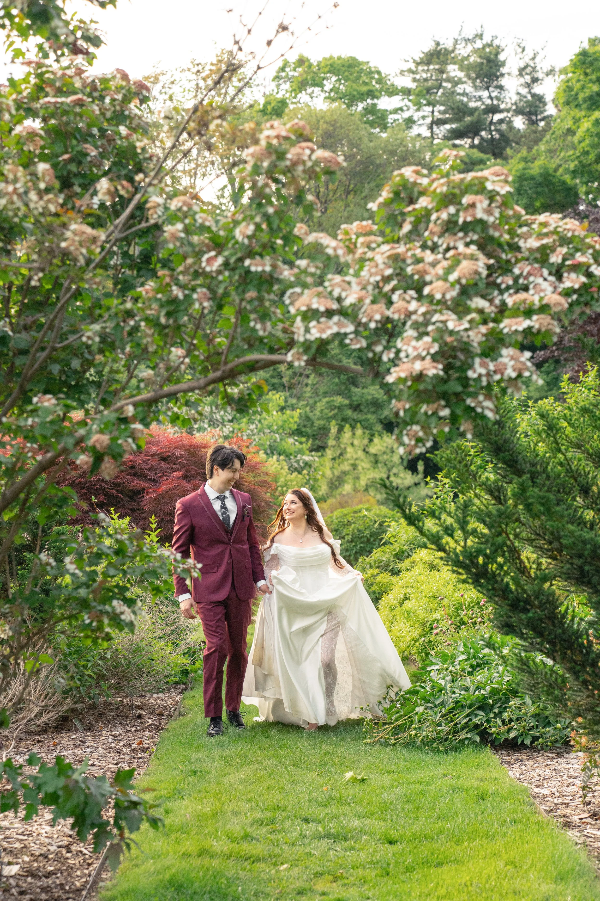 A bride and groom walking through a lush garden, holding hands and smiling at each other, surrounded by green trees and flowering bushes.