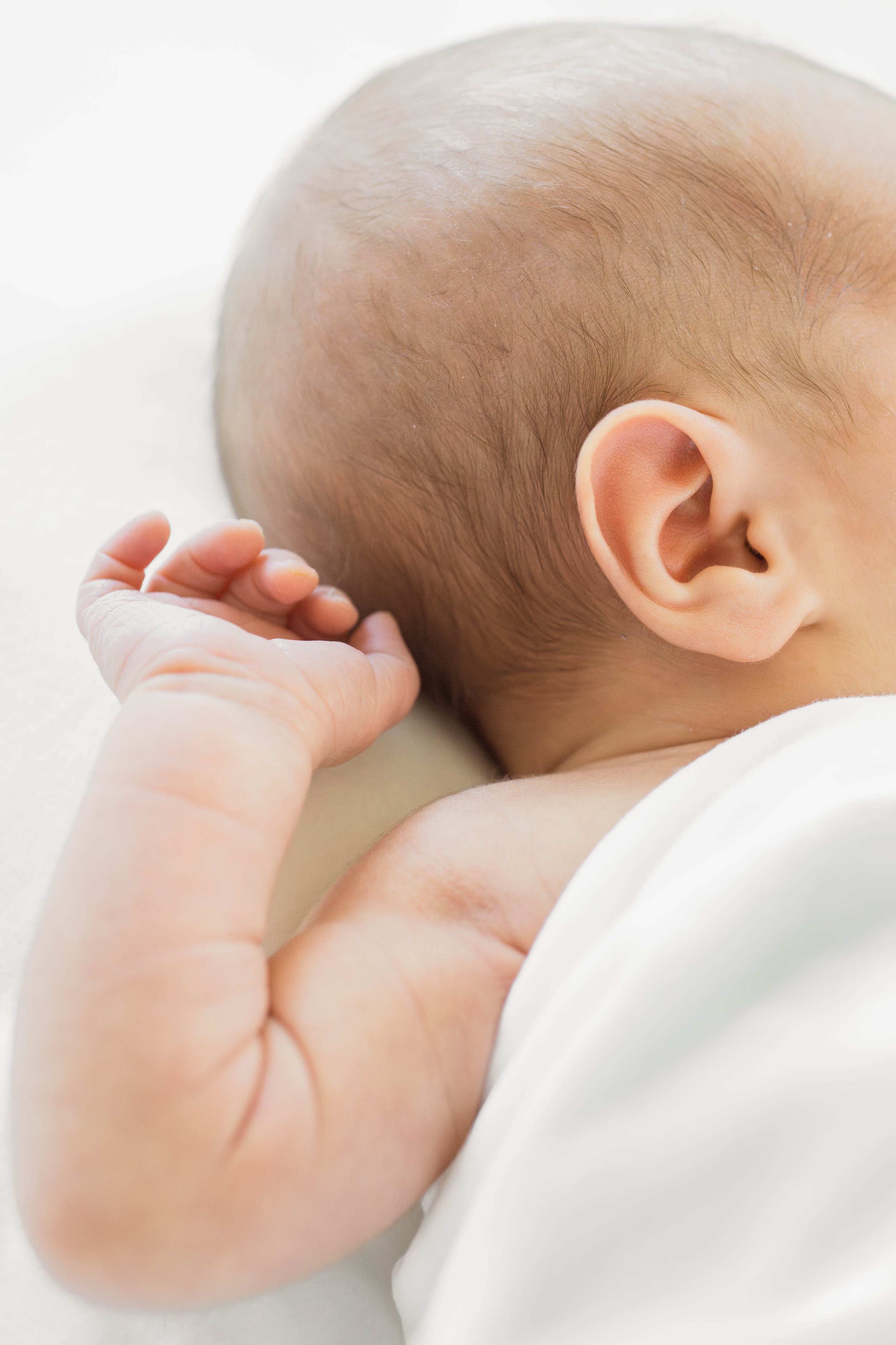 Close-up of a baby's head with a hand resting on the cheek, showing soft skin, baldness, and a small ear, against a white background.