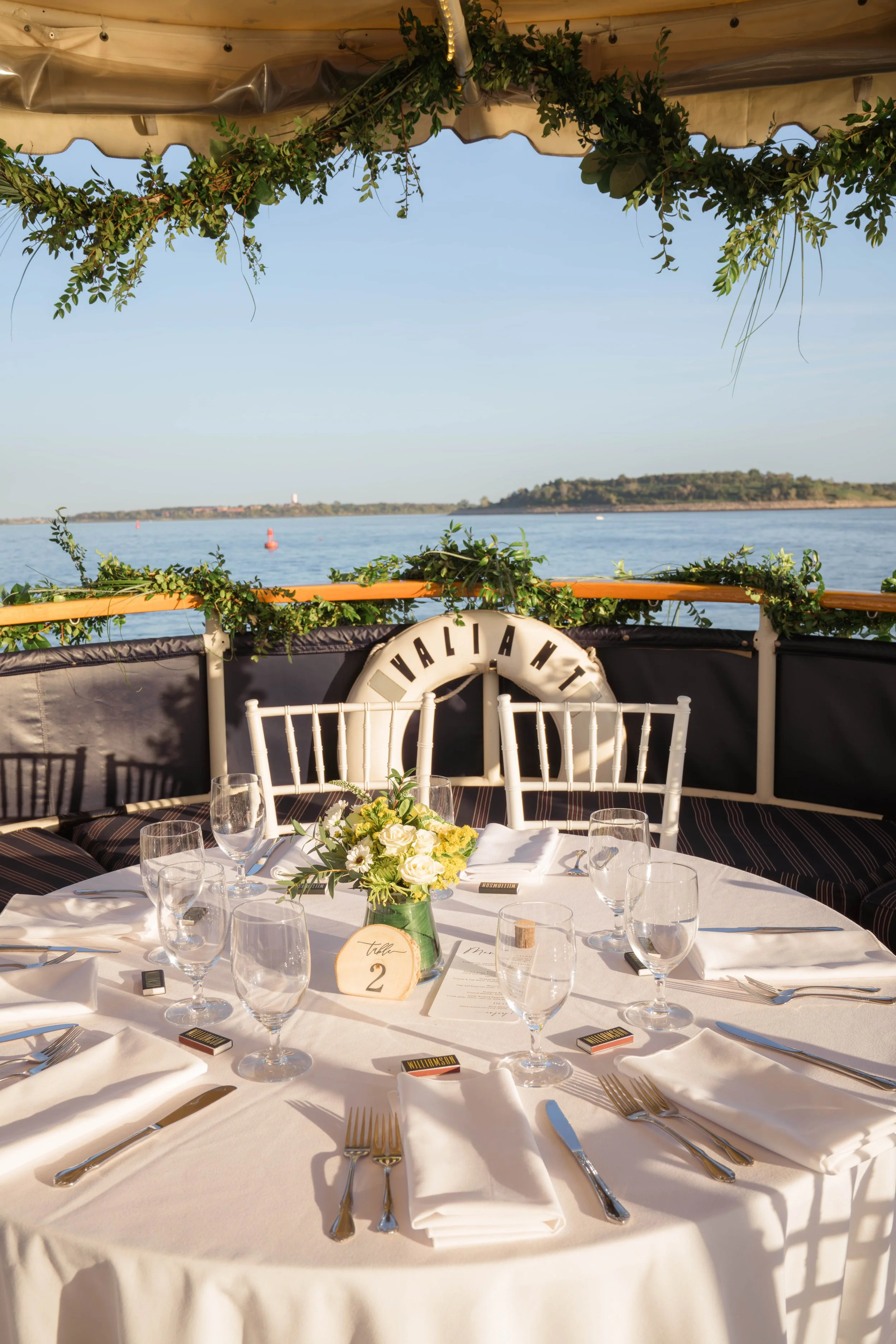A round dining table set for a formal event with wine glasses, silverware, white napkins, and a floral centerpiece, on a boat overlooking a body of water with a distant hill and blue sky in the background.