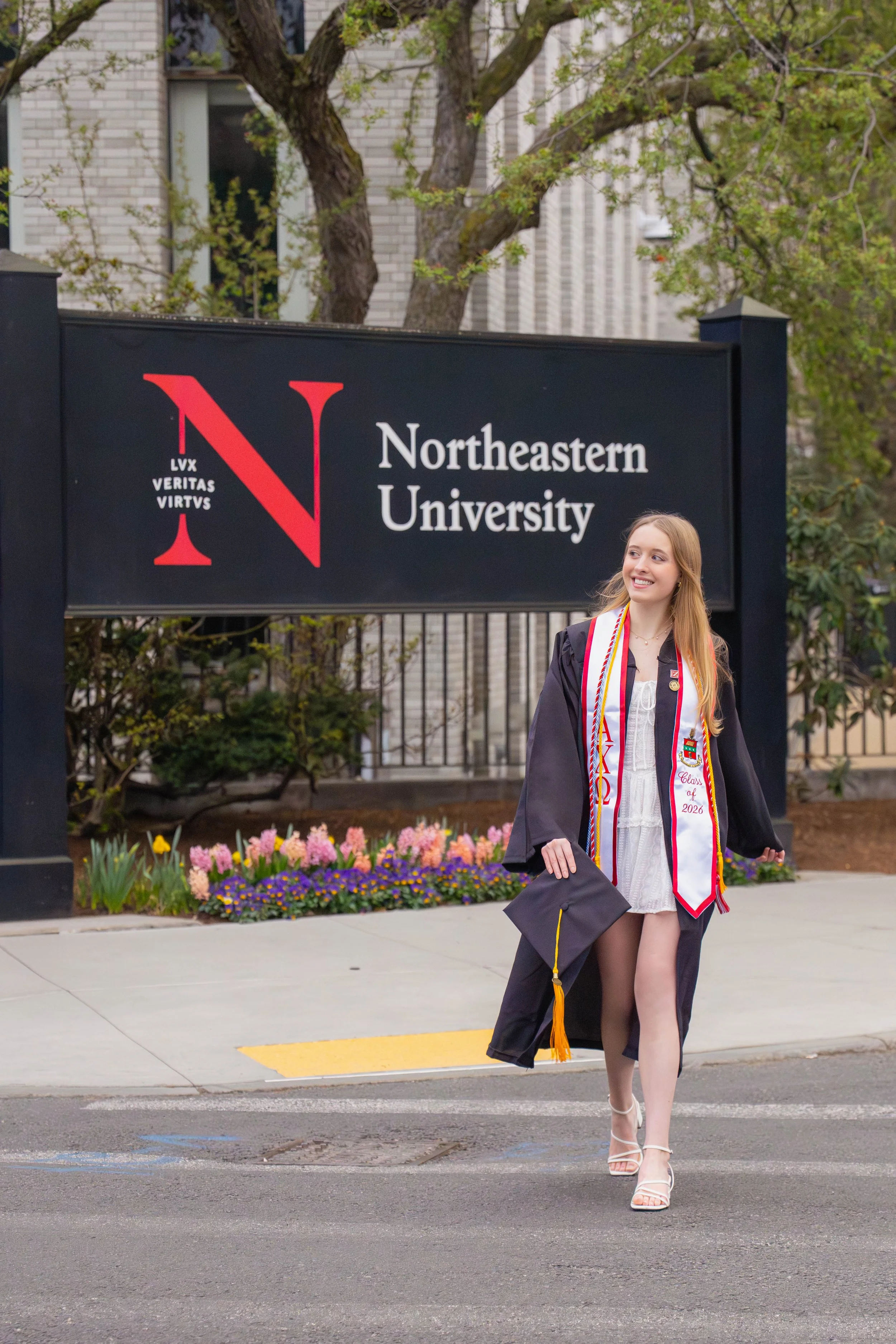 A young woman in a graduation gown and cap walking on a sidewalk in front of a sign for Northeastern University. She is smiling and holding her cap, wearing a white dress underneath her gown, with colorful cords and medals around her neck, and high-h