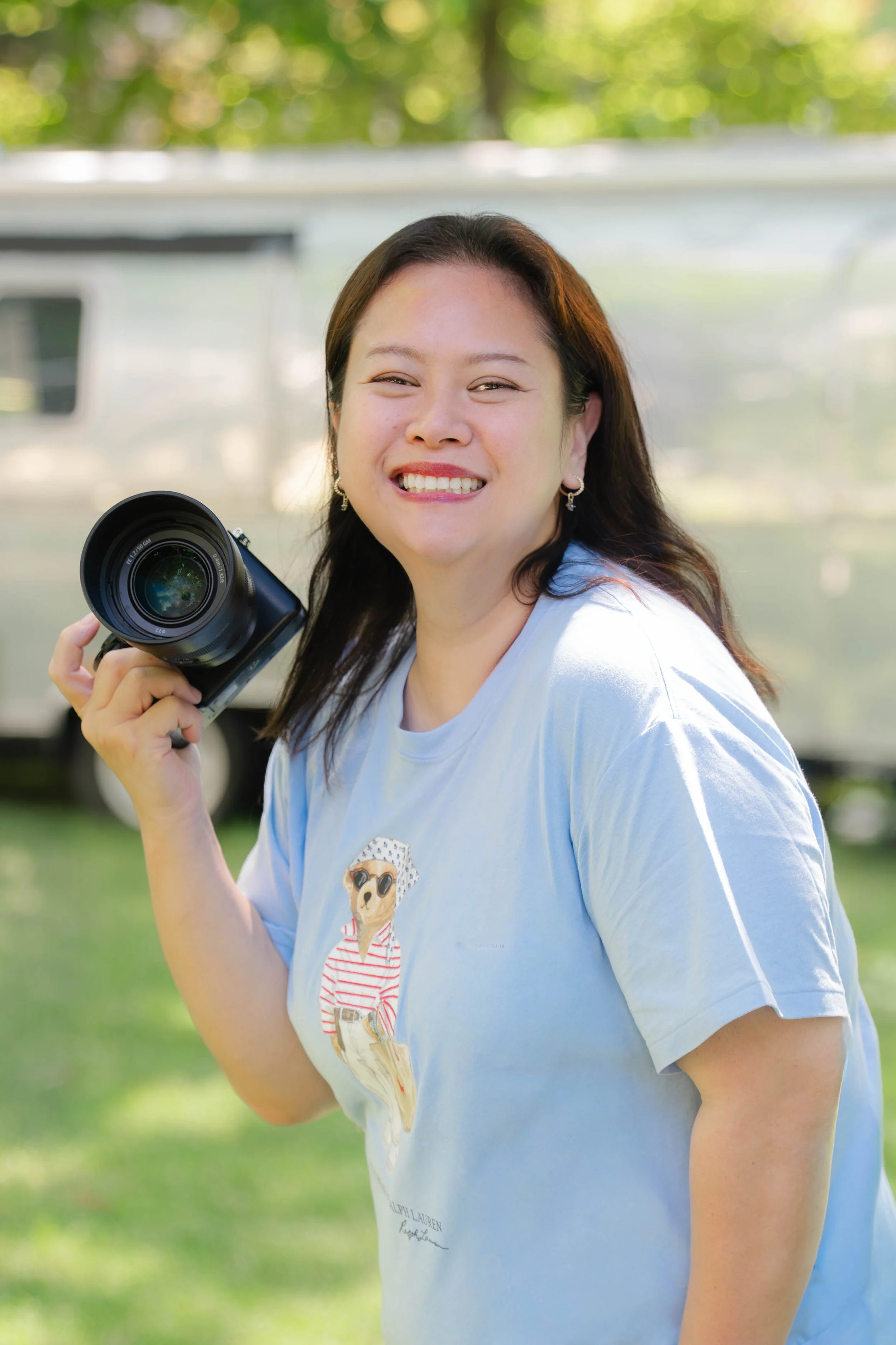 A photo of Mim smiling and holding a camera outdoors with green trees and a vehicle in the background.