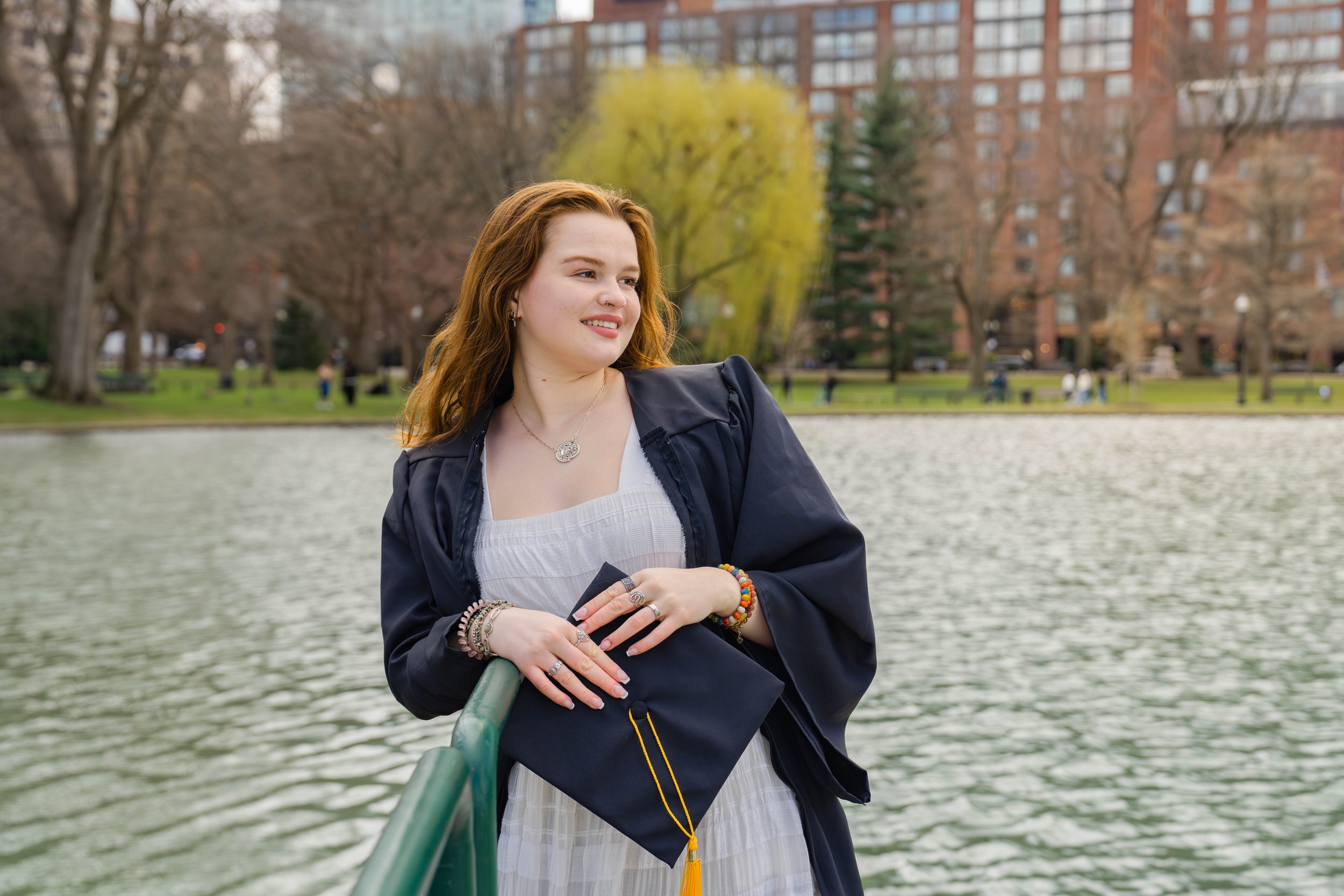 A young woman with red hair standing on a boat near a river, wearing a dark jacket, jewelry, and a light dress, with trees and apartment buildings in the background.