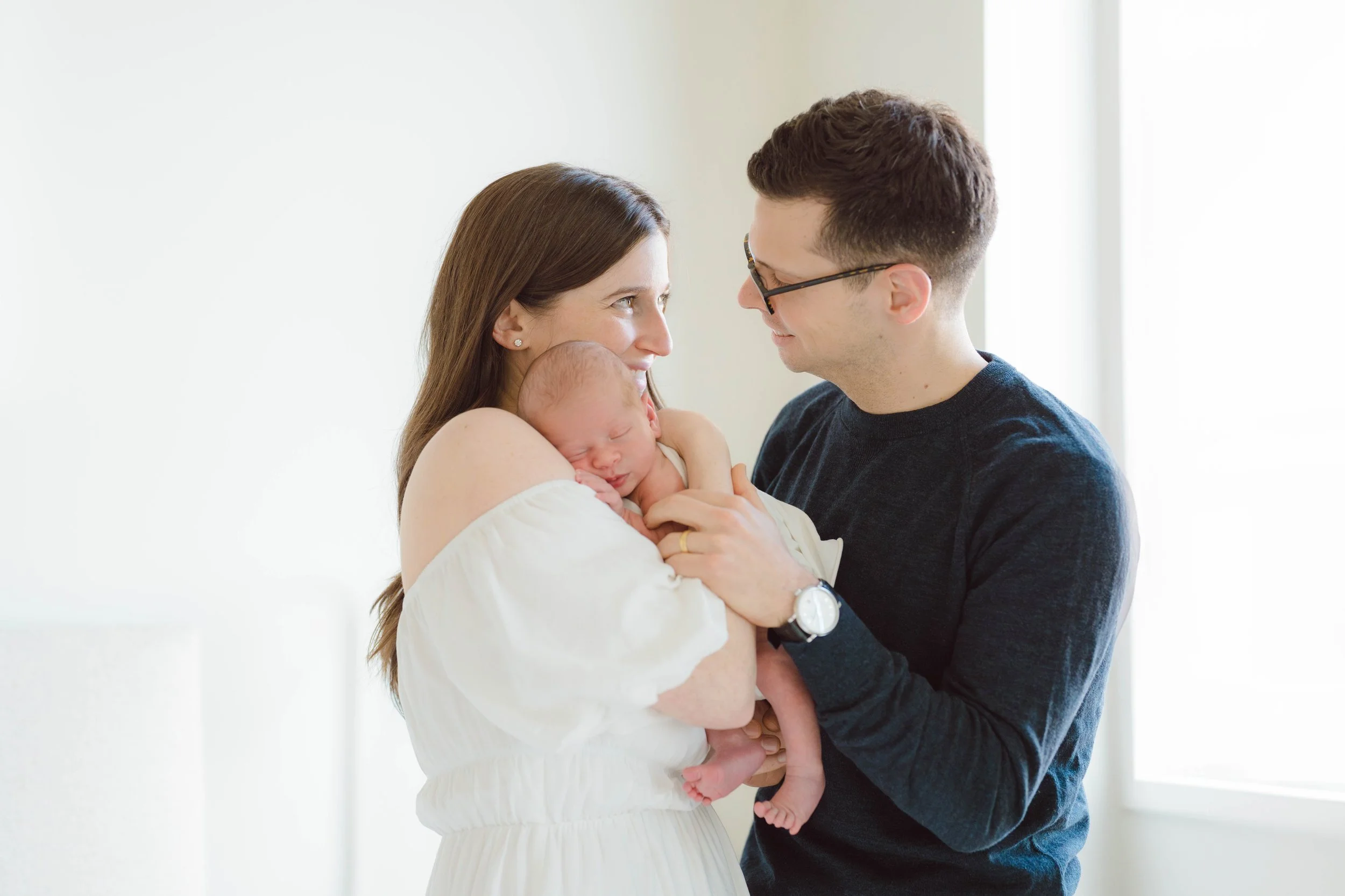 A woman holding a newborn baby, with a man smiling at her, both looking at each other, in a well-lit room.